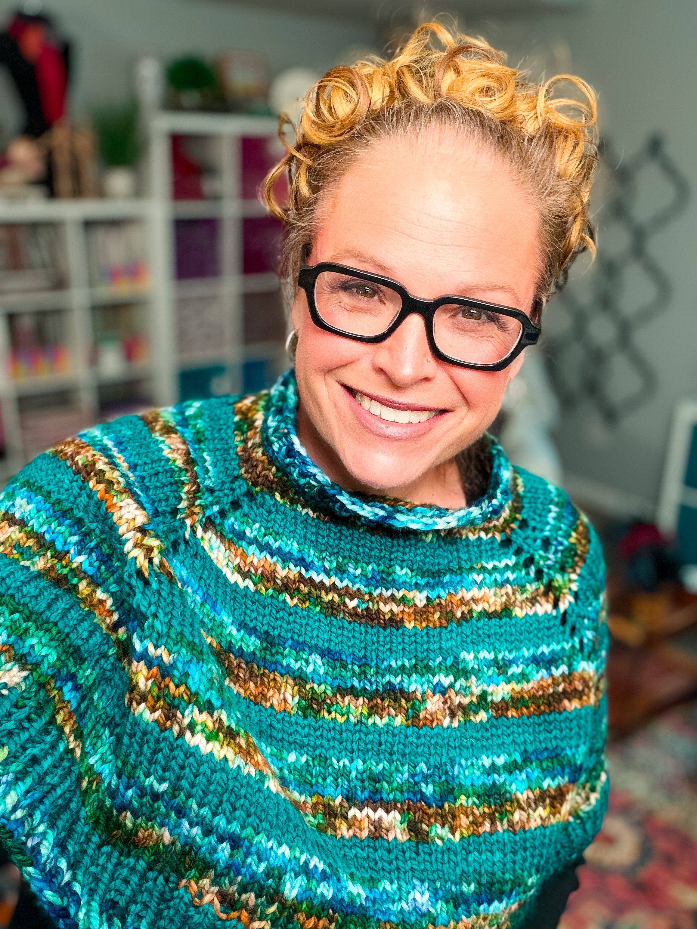 A smiling person with curly blonde hair and glasses is seen wearing the Deschutes Knit Capelet—a chunky, seamless shoulder wrap—in comforting shades of blue, green, and brown. Standing in a cozy, well-lit room filled with shelves and crafts, they showcase the expert craftsmanship of this unique knit capelet & cowl pattern, which adds extra warmth to the inviting atmosphere.