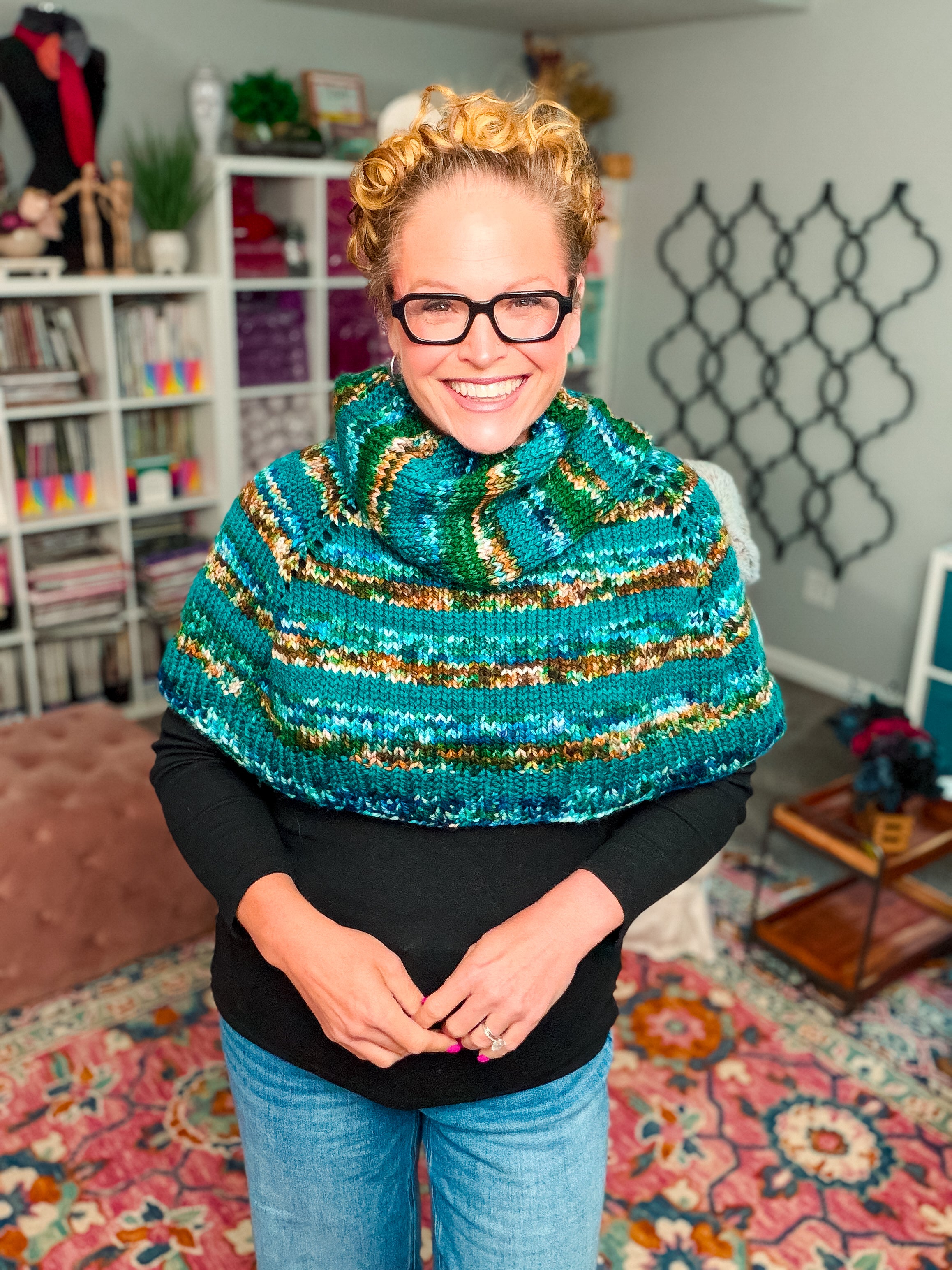 A smiling person with short curly hair and glasses wears the Deschutes Knit Capelet—a chunky knit shoulder wrap featuring the jogless stripe technique—over a black top, standing in a cozy, well-decorated room with bookshelves, yarn, and a patterned rug.