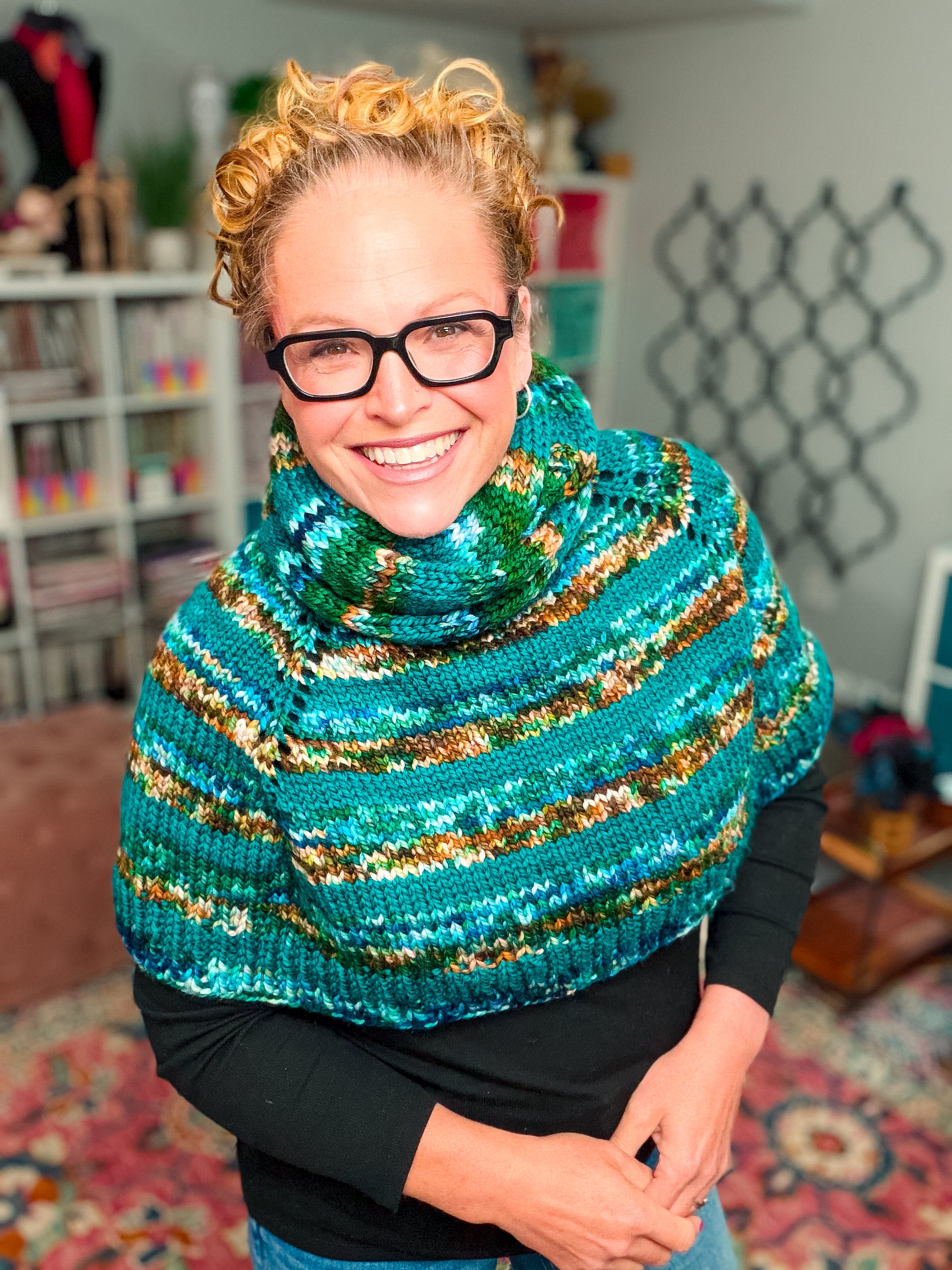 A smiling person with curly blond hair and glasses wears the Deschutes Knit Capelet & Cowl—a chunky, colorful striped shoulder wrap featuring the jogless stripe technique—in a cozy, brightly decorated room with bookshelves and a patterned rug.