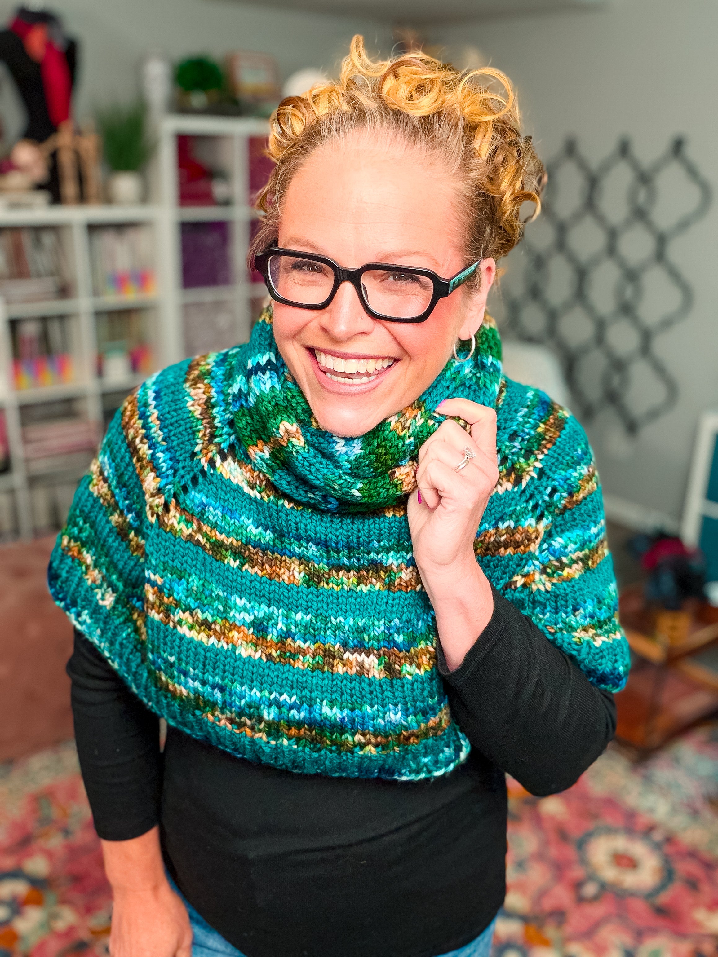 Wearing a colorful, striped chunky knit cowl made from the Deschutes Knit Capelet & Cowl Pattern, a smiling woman with curly blonde hair and glasses stands indoors in a cozy, well-lit room with bookshelves and patterned décor. She pairs the jogless stripe shoulder wrap with a black top.