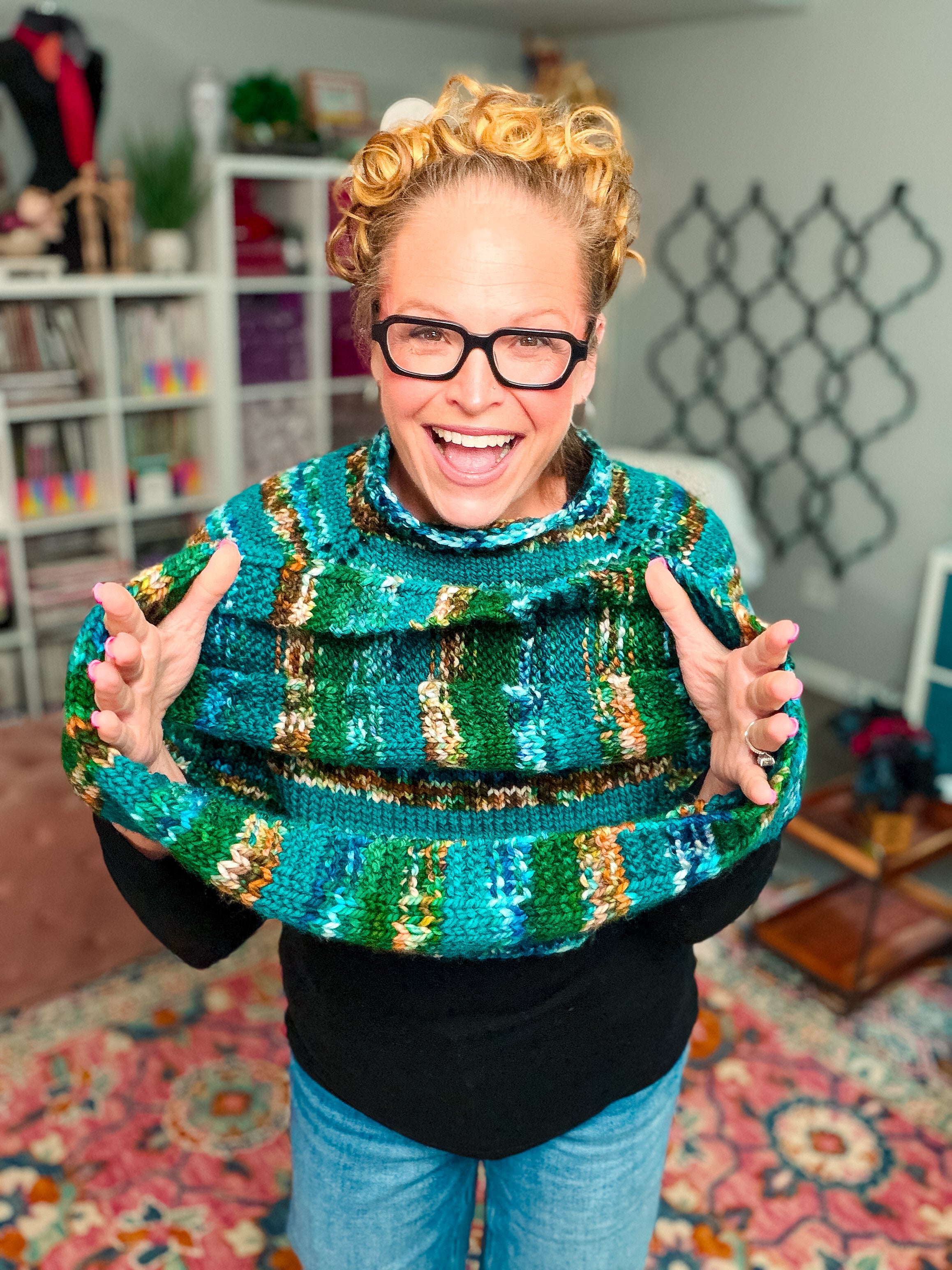 A smiling person with glasses and curly hair wears the Deschutes Knit Capelet & Cowl—a chunky knit shoulder wrap artfully crafted using the jogless stripe technique—over a black shirt, standing in a cozy, colorful room filled with shelves, yarn, and a patterned rug.