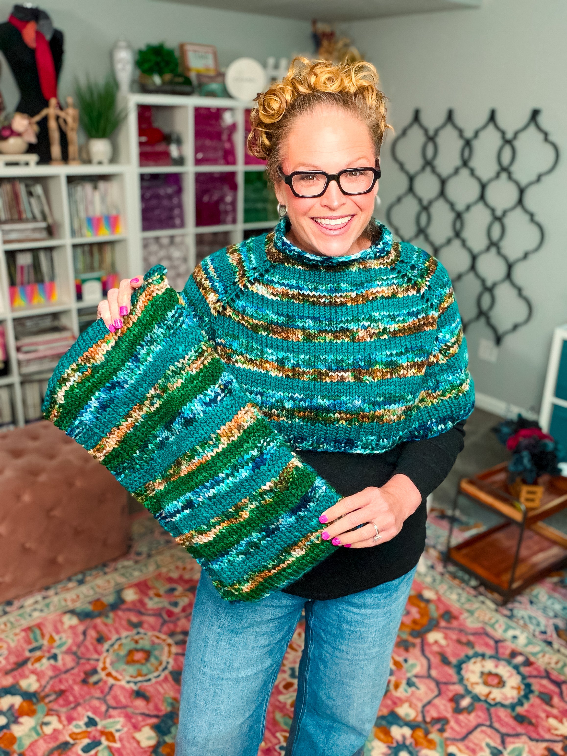 A smiling person with glasses holds a matching green, blue, and brown striped Deschutes Knit Capelet & Cowl—crafted using the jogless stripe technique—standing in a colorful, cozy room filled with craft supplies and vibrant decor.