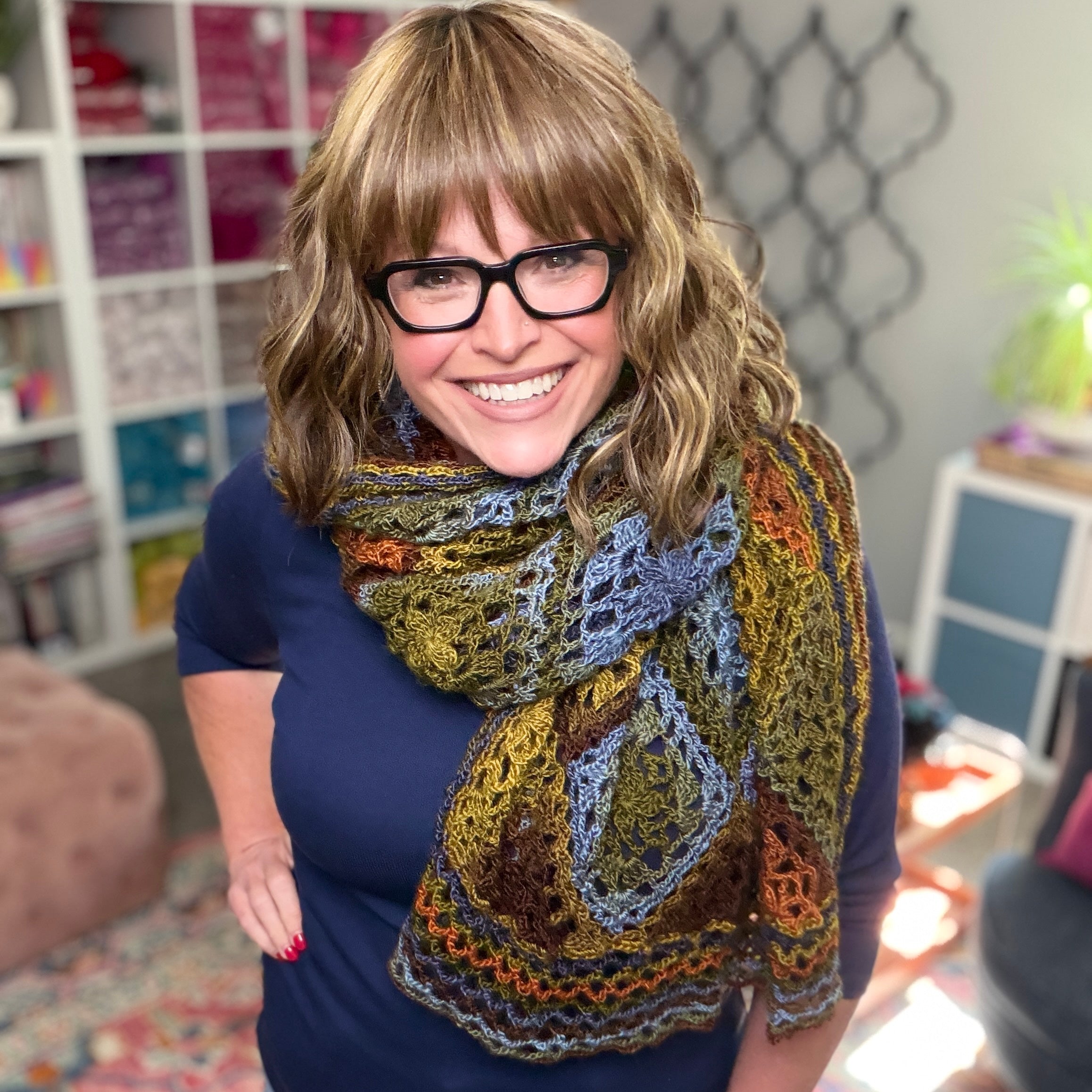 A smiling woman with wavy, light brown hair and glasses wears the Enchanted Crochet Motif Shawl Pattern over a dark blue top. Shelves filled with yarn and a plant appear in the cozy, well-lit background.