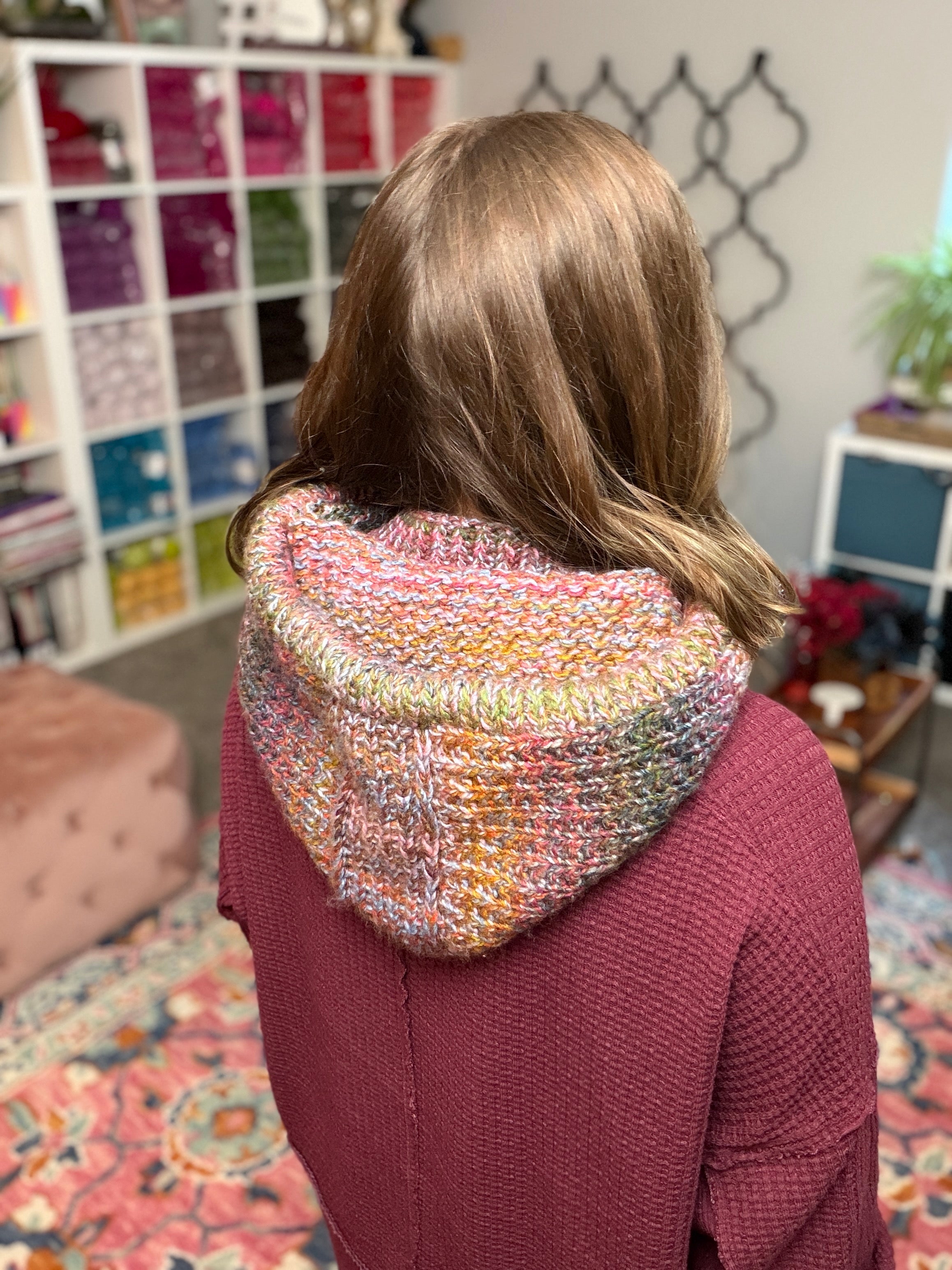 A person models the Cozy Sideline Knit Balaclava - PDF, a multicolored, worsted weight yarn hooded cowl, standing in a cozy room with yarn shelves and vibrant decor in the background.