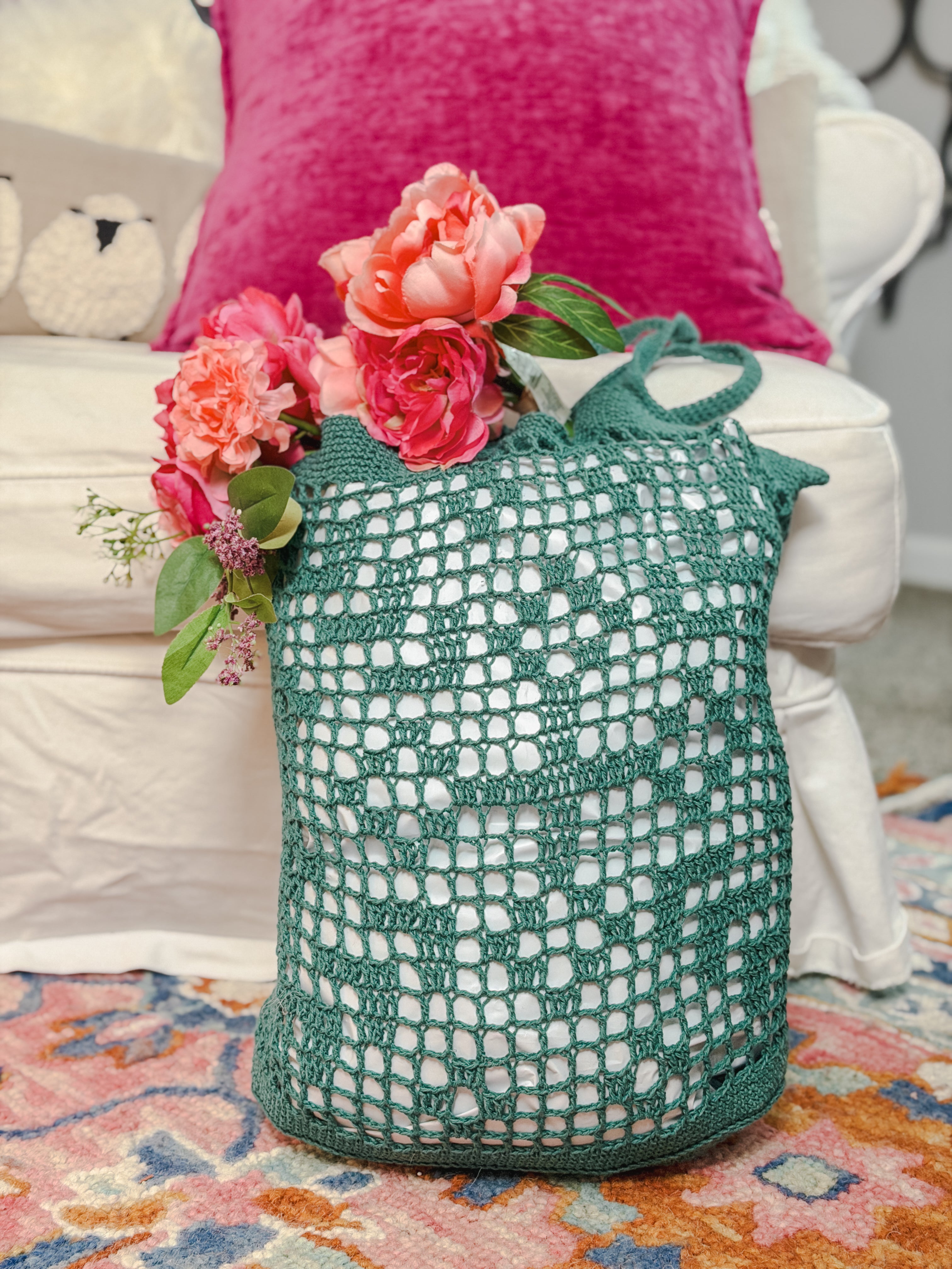 The Farmer’s Market Filet Crochet Bag, a reusable mesh tote, holds pink flowers and sits on a colorful rug in front of a white sofa with a bright pink pillow.