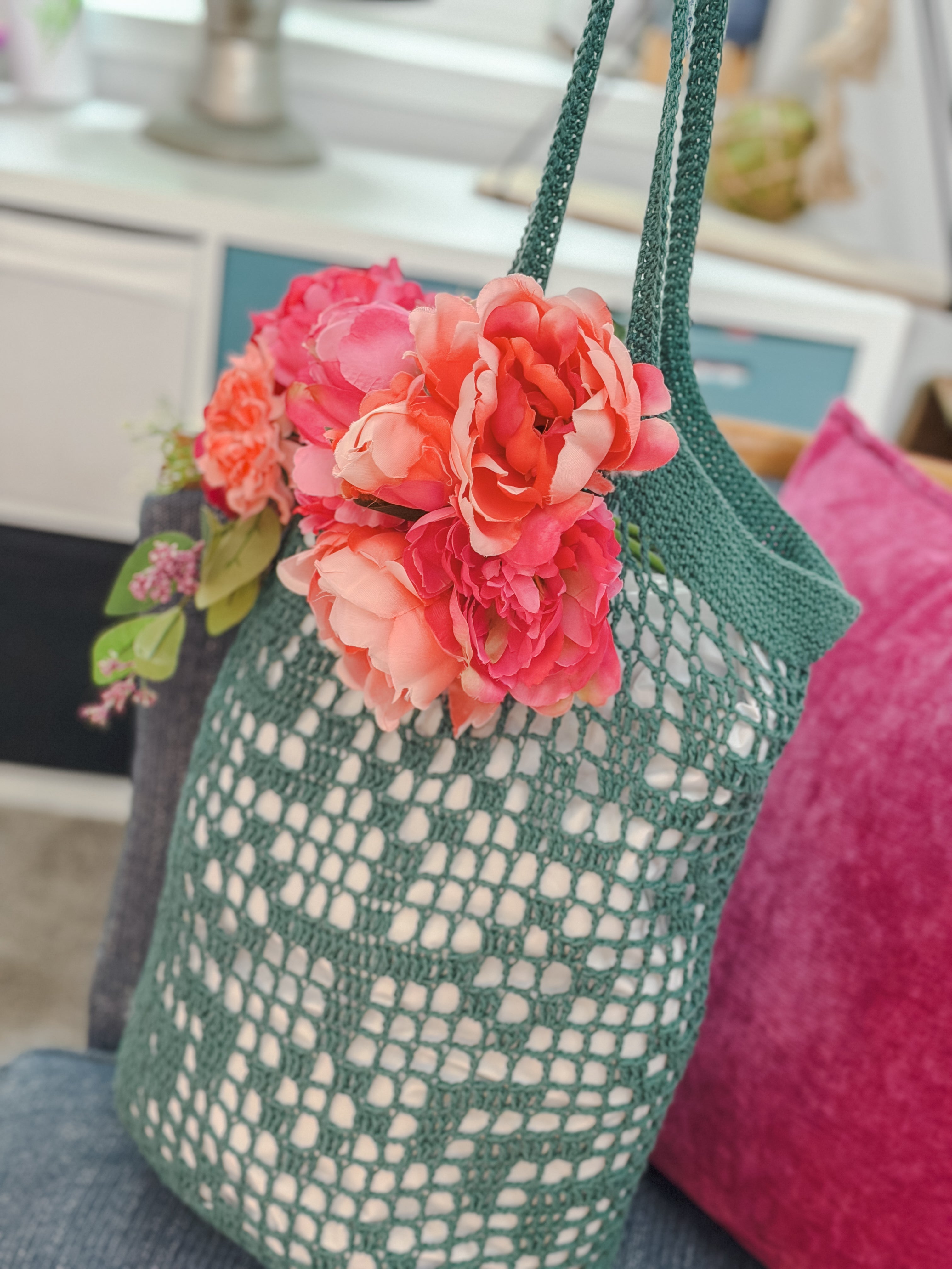 The Farmer’s Market Filet Crochet Bag—a green mesh tote decorated with bold pink and coral faux flowers—rests on a chair with a magenta pillow in a cozy indoor space.