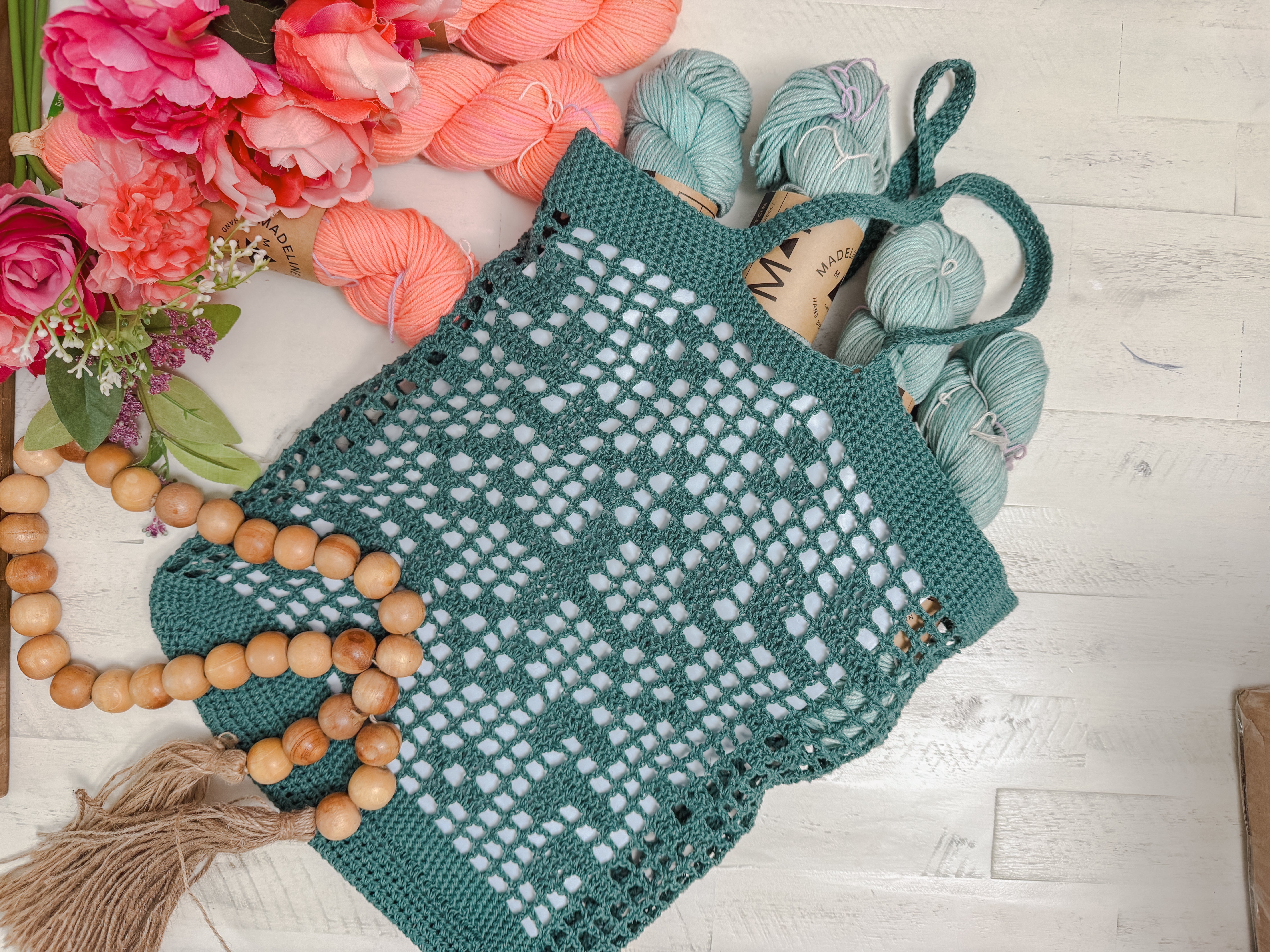 The Farmer’s Market Filet Crochet Bag Pattern shows a reusable green mesh tote filled with blue yarn skeins, placed on a light wooden surface beside pink flowers, orange yarn, and a long beaded tassel garland.