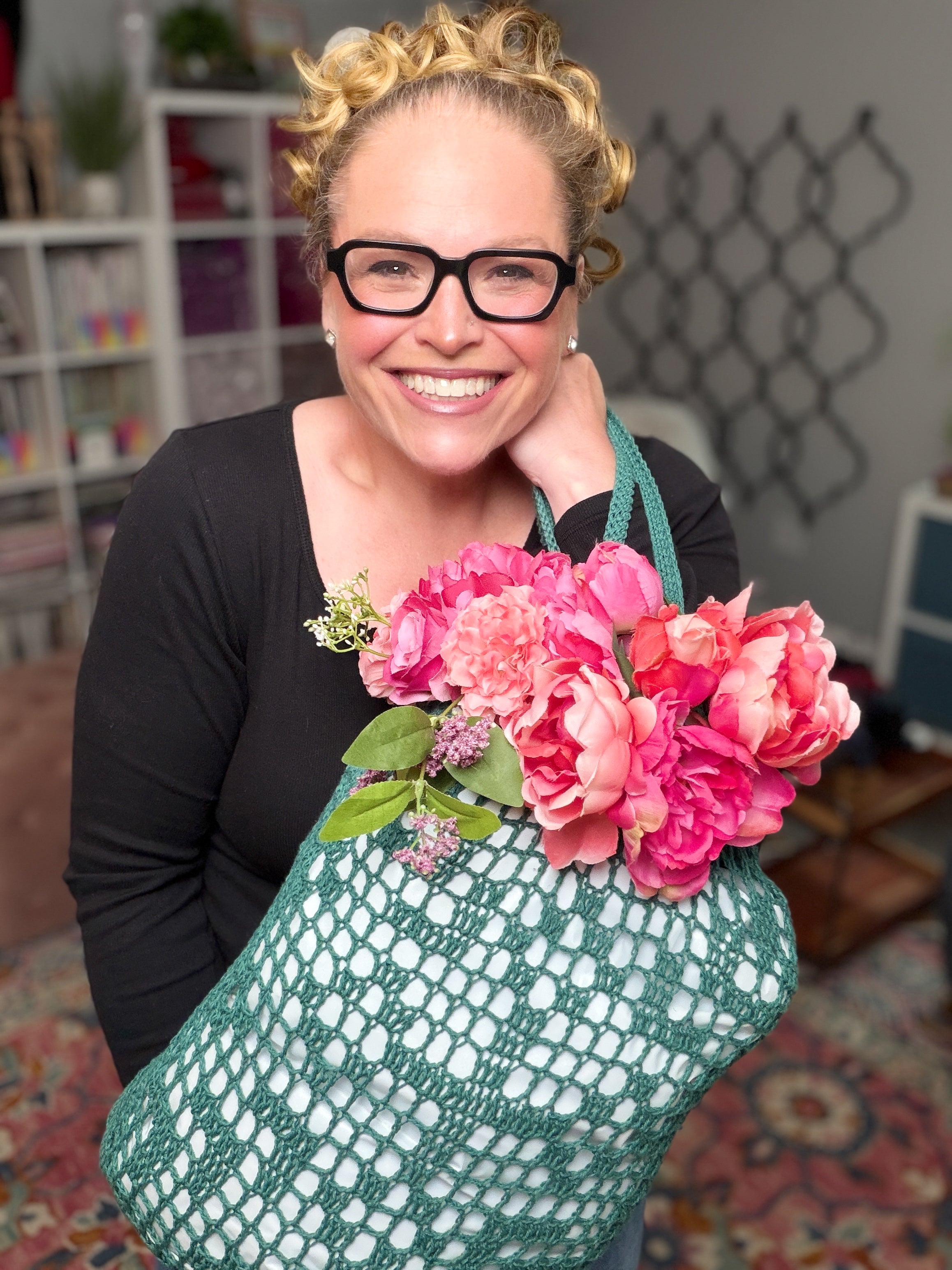 A smiling person with glasses holds the Farmer’s Market Filet Crochet Bag—a reusable mesh tote—filled with pink flowers. Behind them are shelves and a patterned rug.