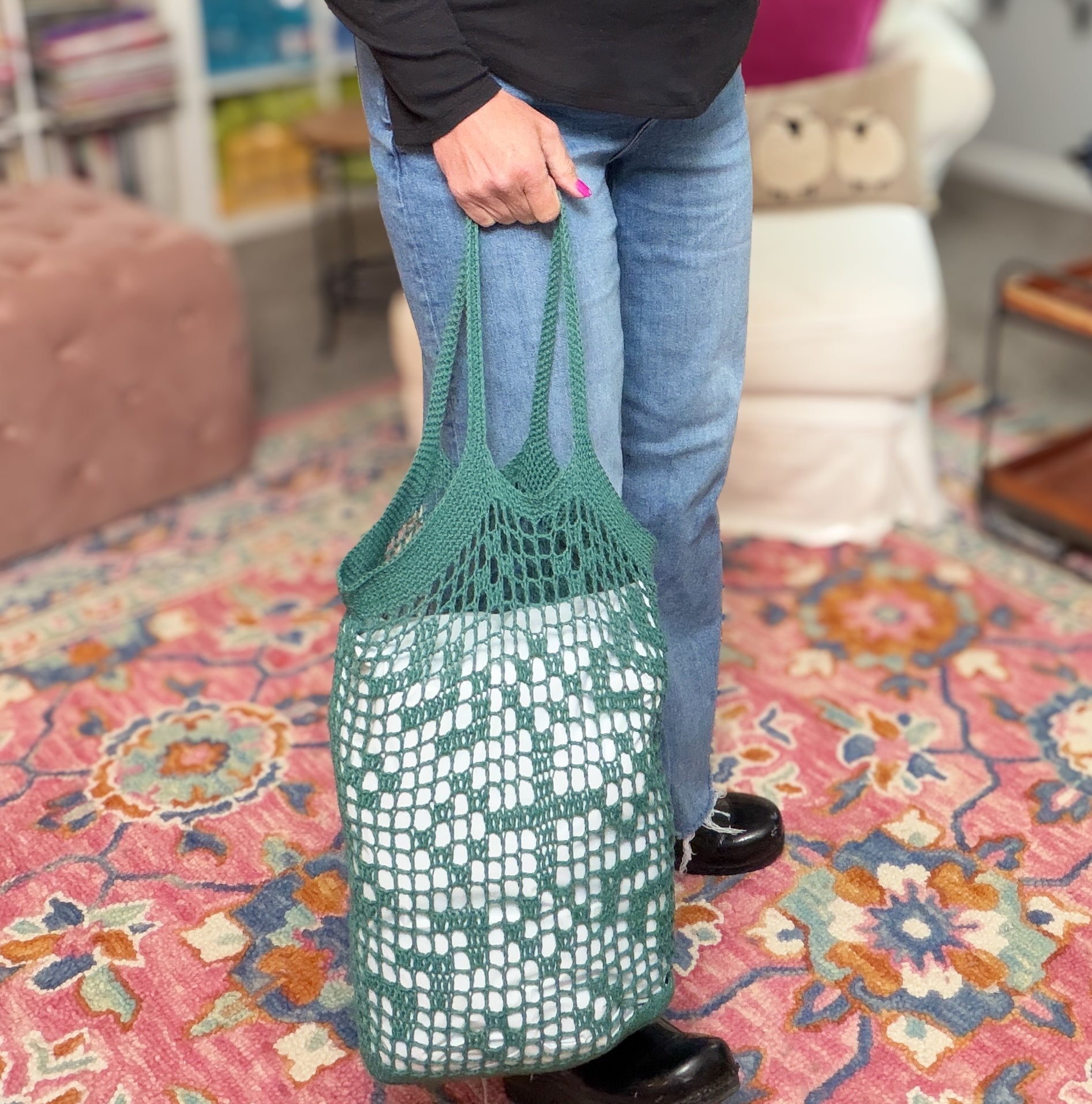 Wearing jeans and black shoes, a person holds the Farmer’s Market Filet Crochet Bag—a reusable mesh tote with a patterned interior—while standing on a colorful rug in a cozy room with a white chair and furnishings.