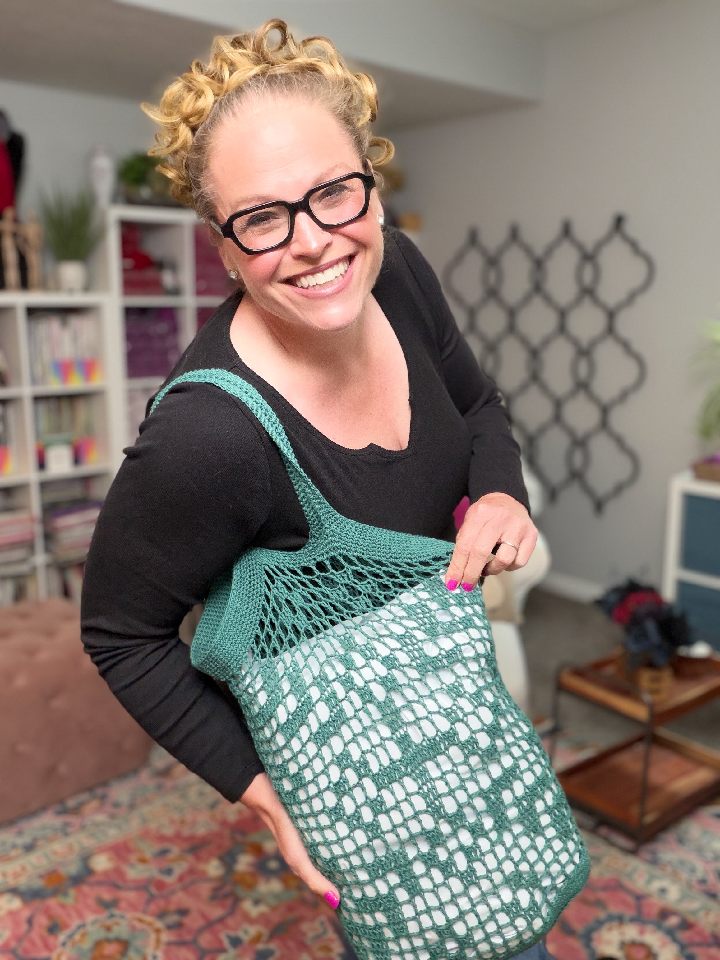 A smiling woman with curly blonde hair and glasses holds the Farmer’s Market Filet Crochet Bag – a reusable mesh tote – in a colorful room with patterned rugs and shelves in the background.