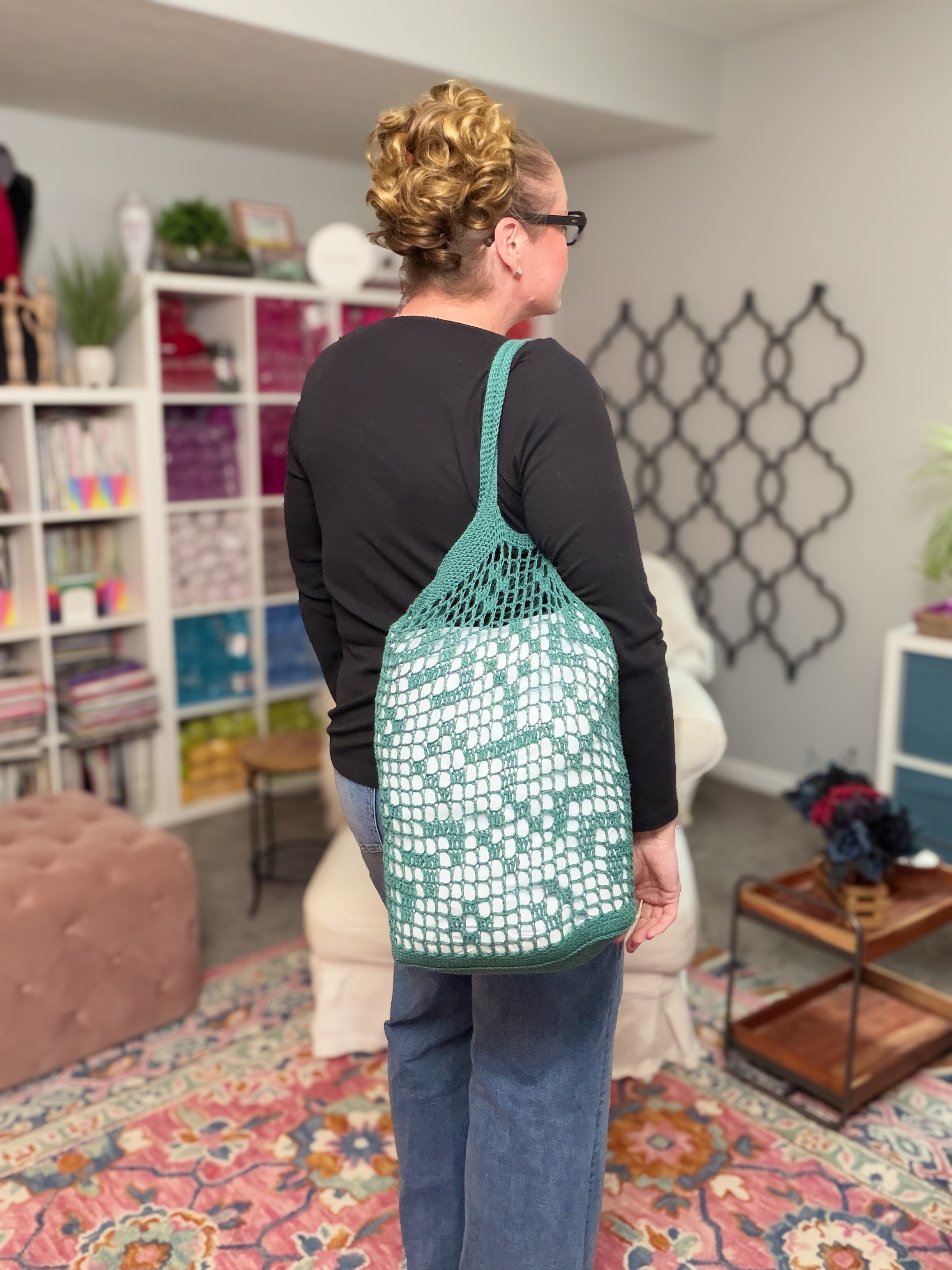 A woman with blonde curly hair in a bun, wearing glasses and a black top, stands indoors with her back to the camera, carrying the Farmer’s Market Filet Crochet Bag—a reusable mesh tote. Shelves with yarn and decor are in the background.