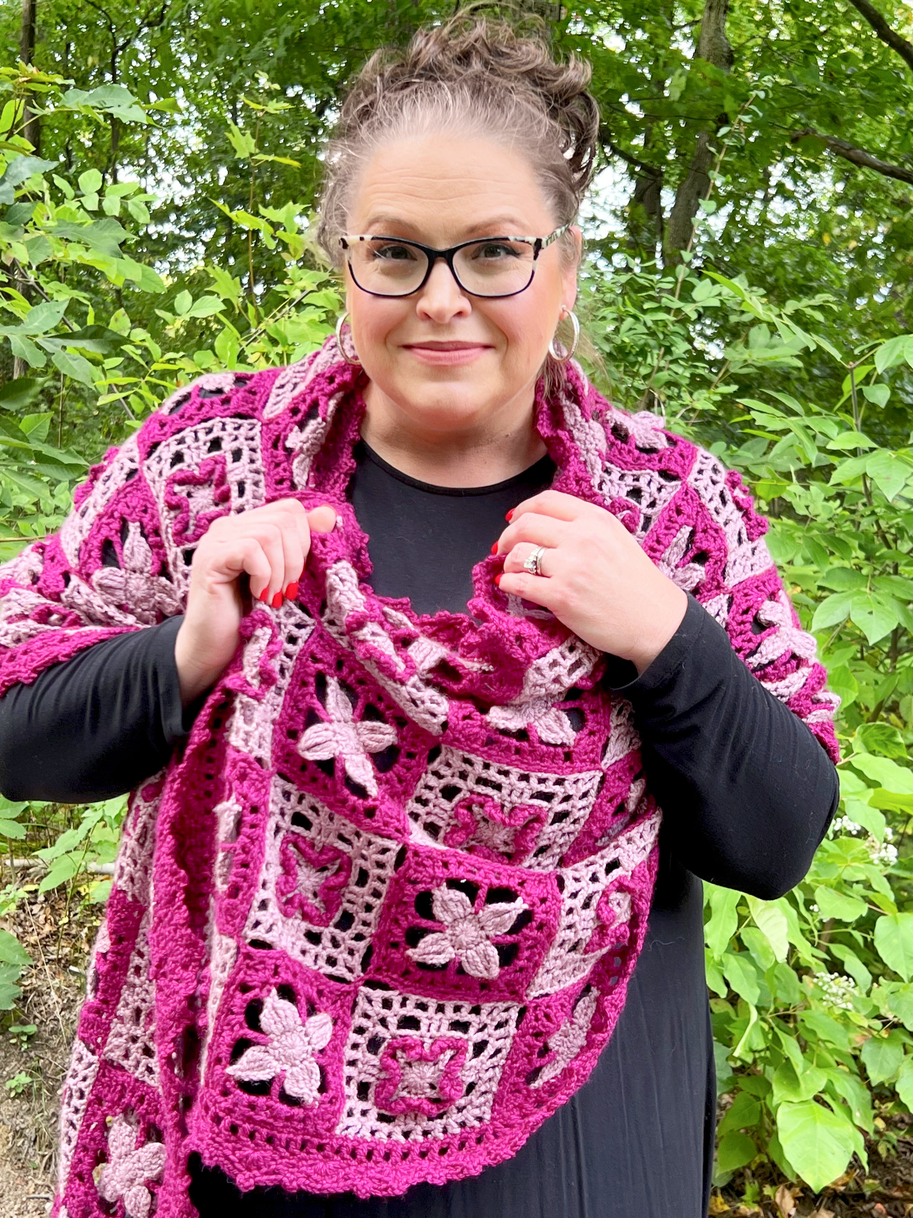Person wearing a pink and purple crocheted shawl outdoors with greenery in the background