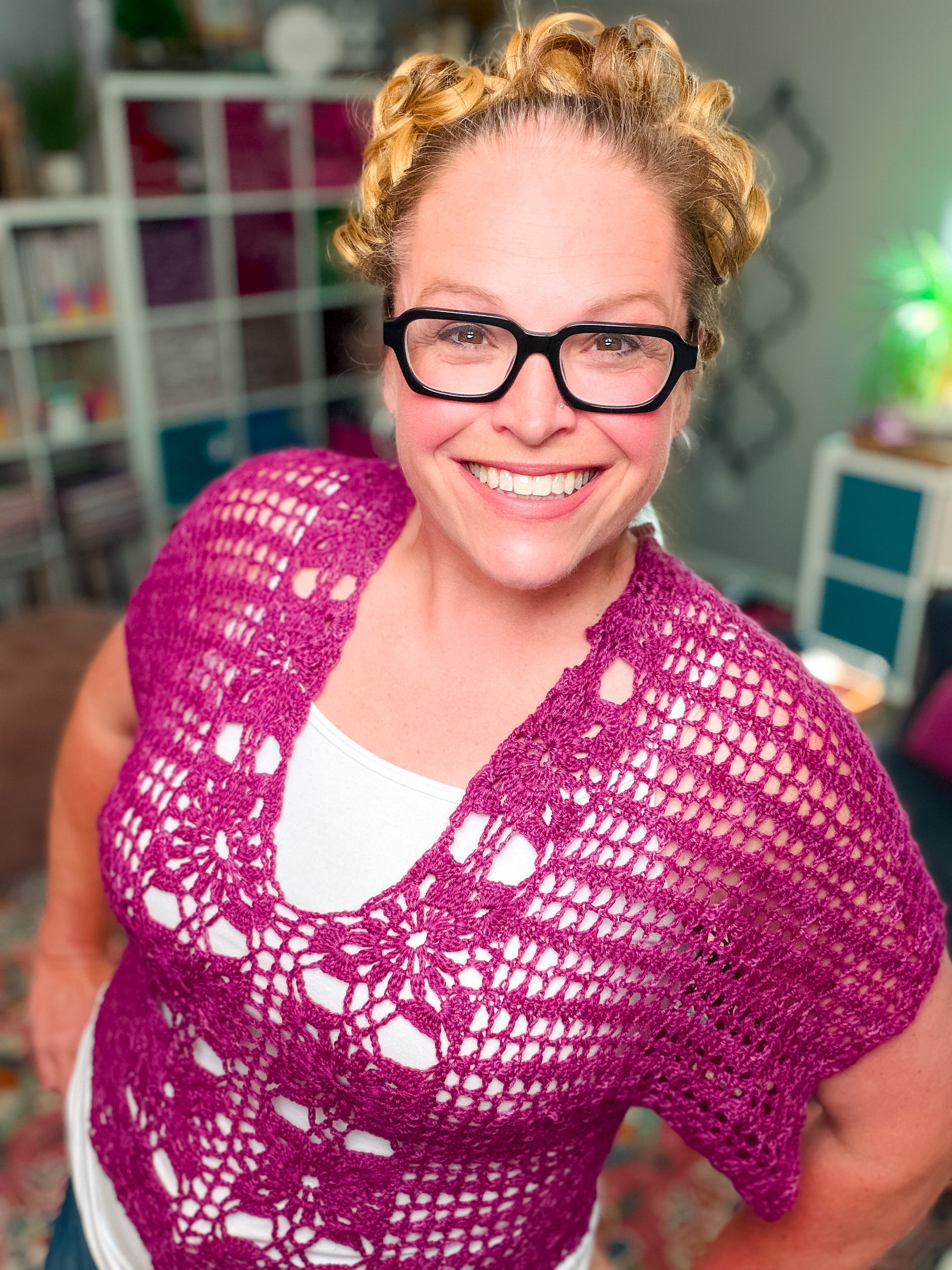 A smiling blonde woman in black glasses and a white top layers the Floral Motif Crochet Summer Tee (PDF pattern, sizes S–5X) in a cozy, colorful room with patterned rug and shelves.