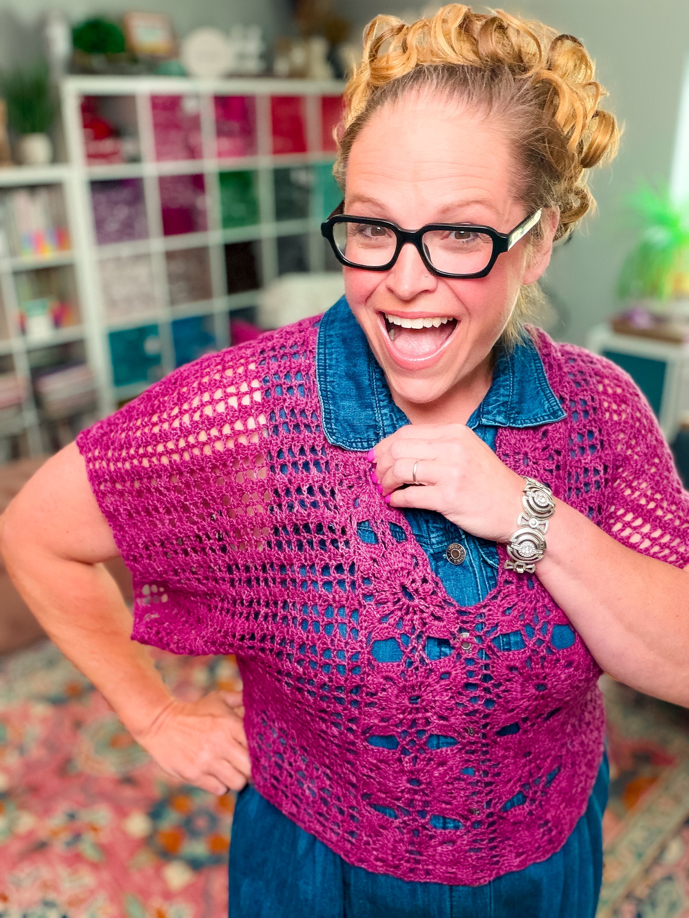 A smiling woman with blonde hair and glasses models the Floral Motif Crochet Summer Tee (PDF pattern, S–5X), a purple lace crochet top, over a blue dress. She stands in front of colorful yarn shelves, posing confidently with one hand on her hip.