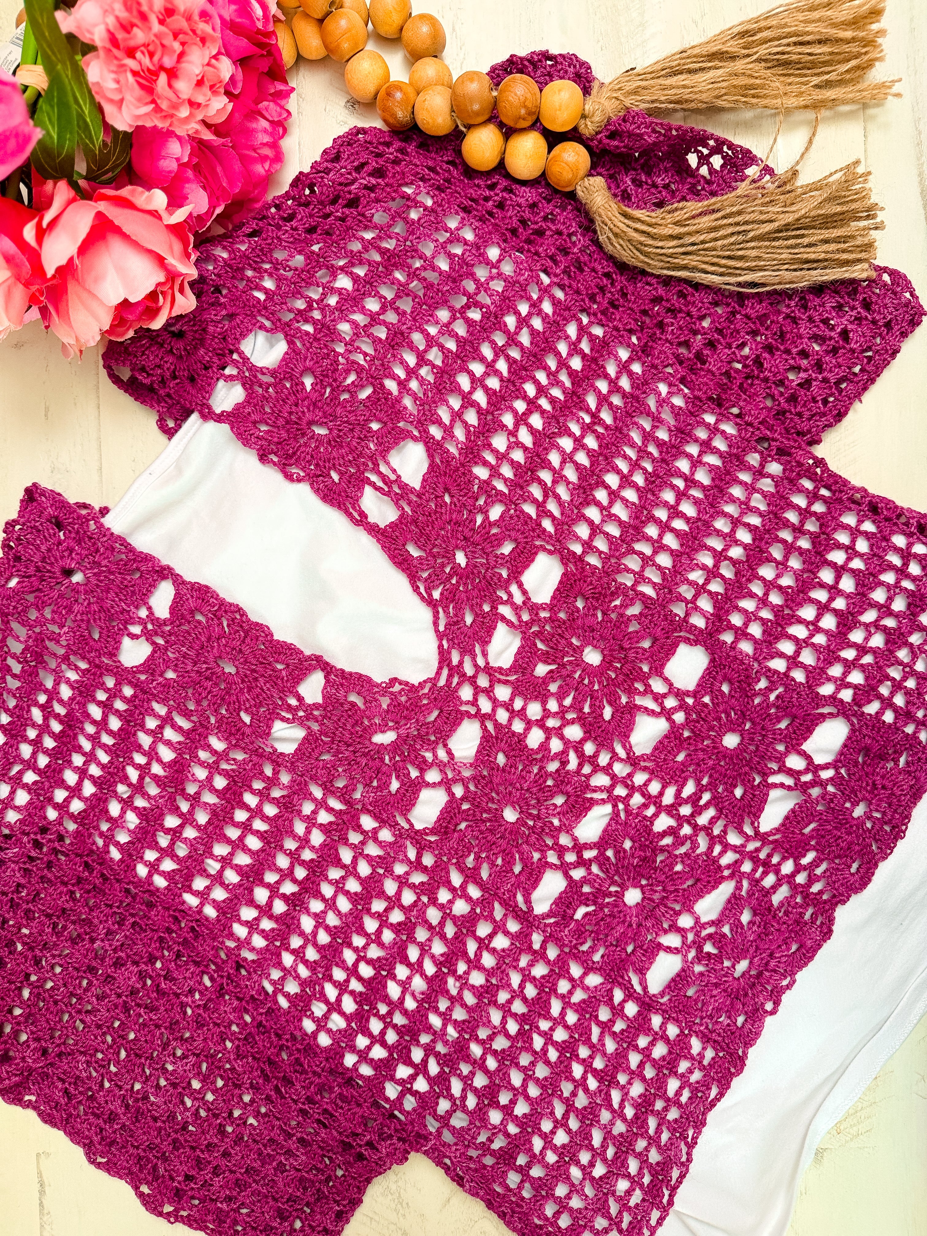 The Floral Motif Crochet Summer Tee Pattern – Lace Crochet Top PDF (Sizes S–5X) in magenta is displayed on a white surface beside pink flowers and a beaded tassel accessory.