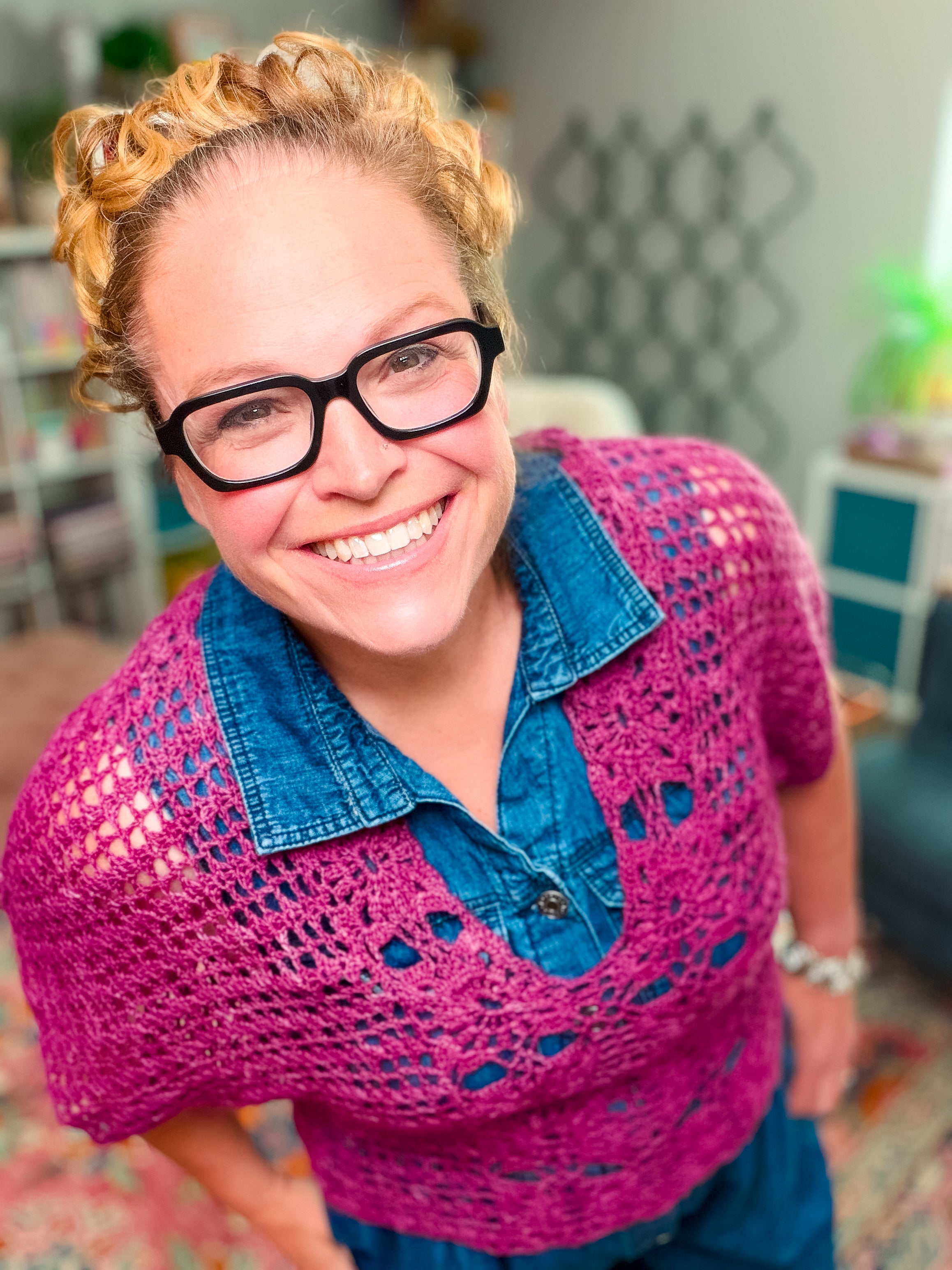A smiling person with curly blond hair, black glasses, and a blue denim shirt models the Floral Motif Crochet Summer Tee Pattern – a pink lace crochet top (PDF, sizes S–5X), indoors with blurred shelves and decor in the background.