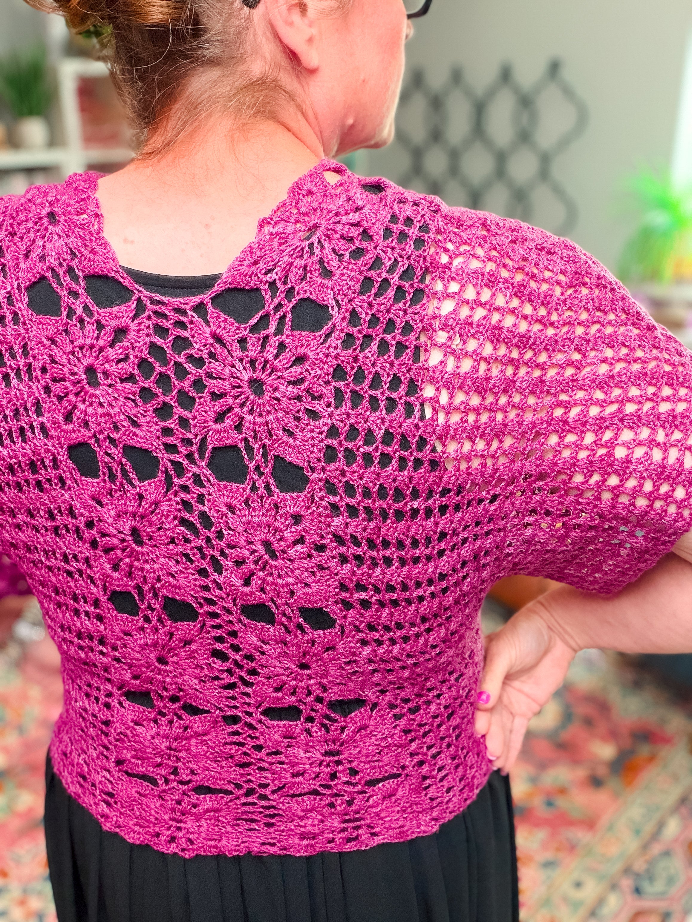 A person with hair in a bun models the Floral Motif Crochet Summer Tee Pattern – a bright pink lace crochet top (PDF, sizes S–5X) with short sleeves and intricate floral patterns, styled over a black shirt in a softly blurred indoor photo.