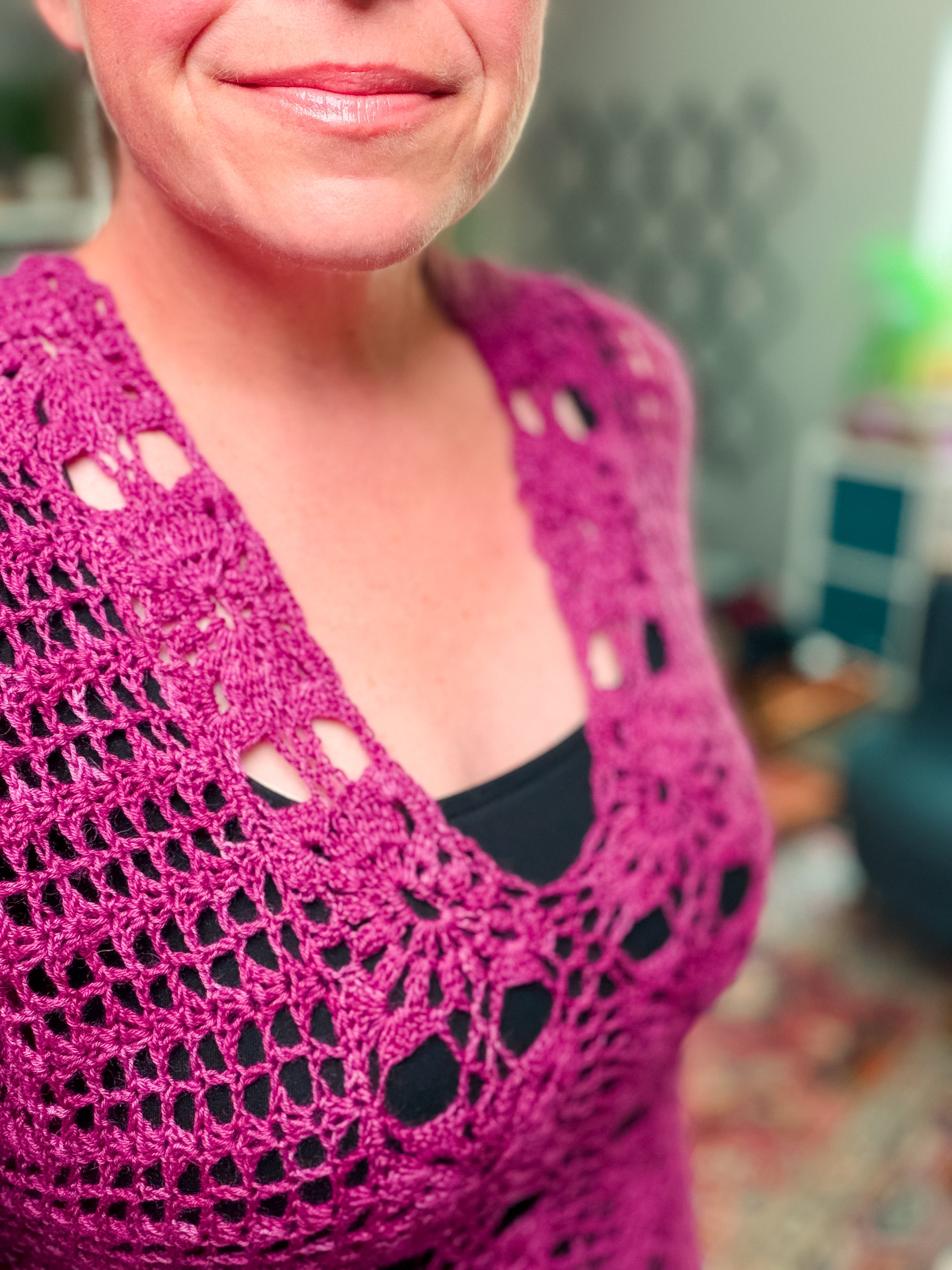 A woman wears the Floral Motif Crochet Summer Tee, a lace crochet top (PDF pattern, sizes S–5X), over a black tank, smiling slightly. The focus is on the detailed design; the softly lit room background is blurred.