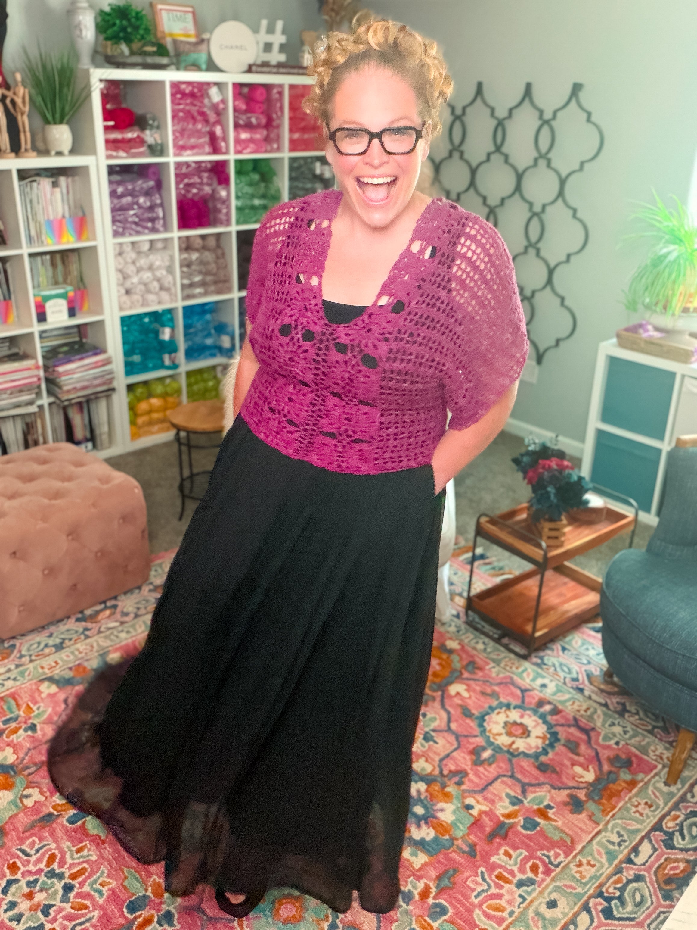 A smiling woman with curly blonde hair and glasses wears the Floral Motif Crochet Summer Tee, a lace crochet top. She stands in a colorful, cozy room with yarn shelves, plants, and patterned rugs.