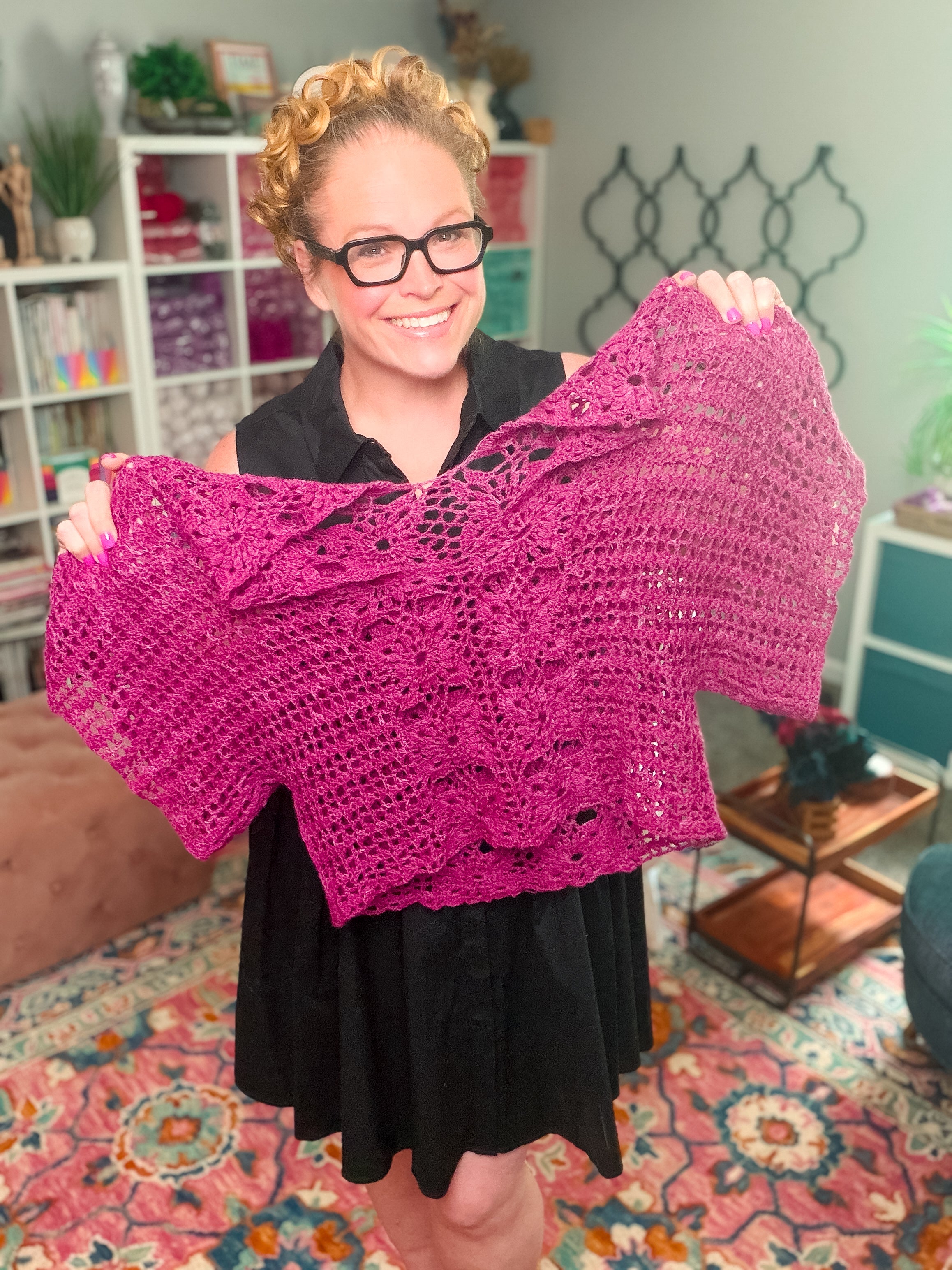 A smiling person wearing glasses and a black dress holds up the Floral Motif Crochet Summer Tee—a purple lace crochet top—inside a brightly lit, colorful room with shelves, plants, and patterned decor. Available as a PDF pattern in sizes S–5X.