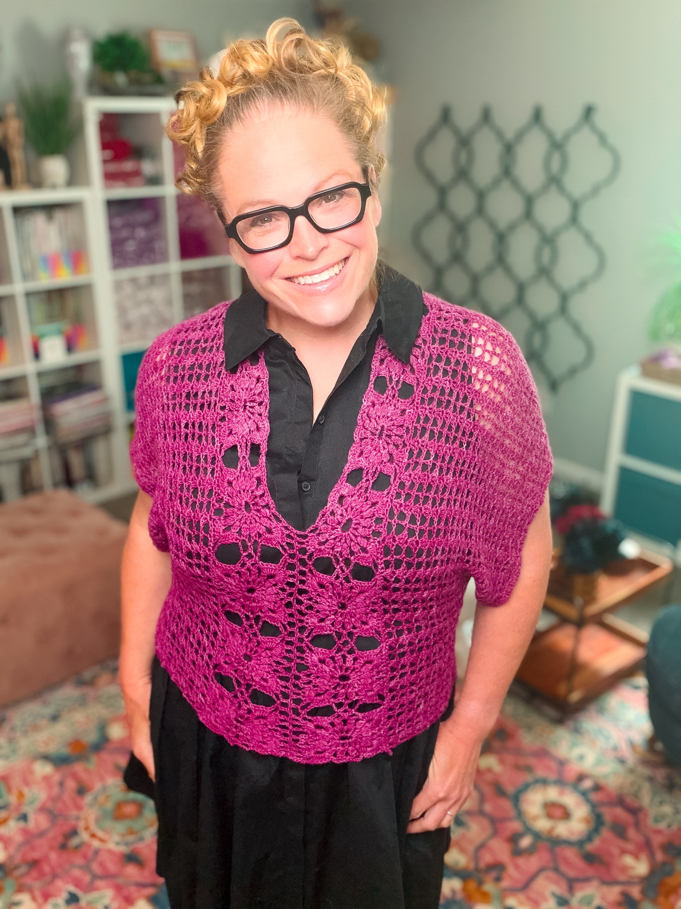 A smiling woman with curly blonde hair and glasses wears a black dress layered with the Floral Motif Crochet Summer Tee—a purple lace crochet top. She stands in a cozy, colorful room decorated with books, plants, and patterned decor.