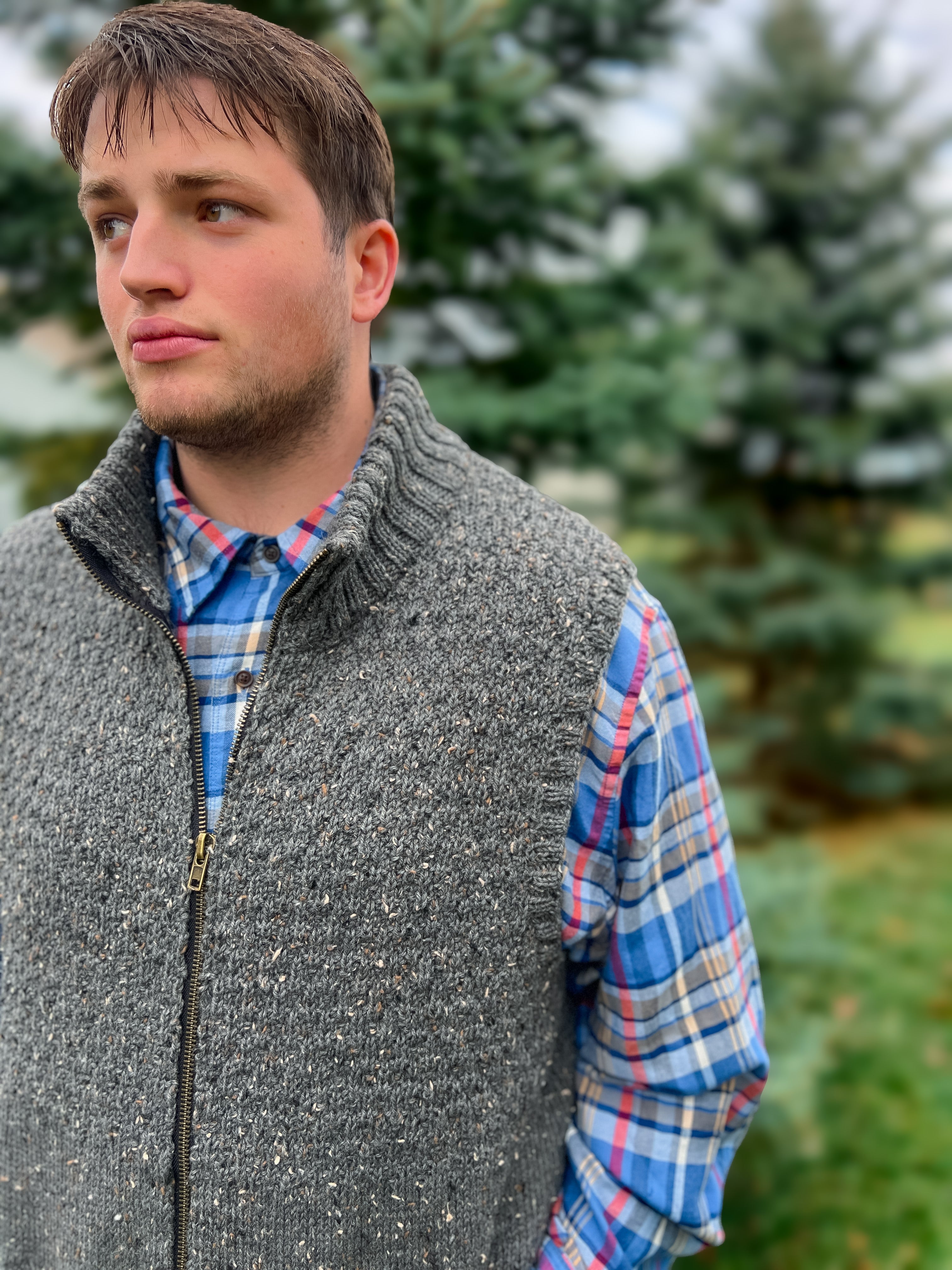 A young man stands outdoors in front of pine trees, gazing thoughtfully to the left while wearing the Harbor Ridge Knit Vest layered over a blue plaid shirt—an ensemble that showcases the timeless appeal of the Harbor Ridge Knit Vest & Sweater Pattern.