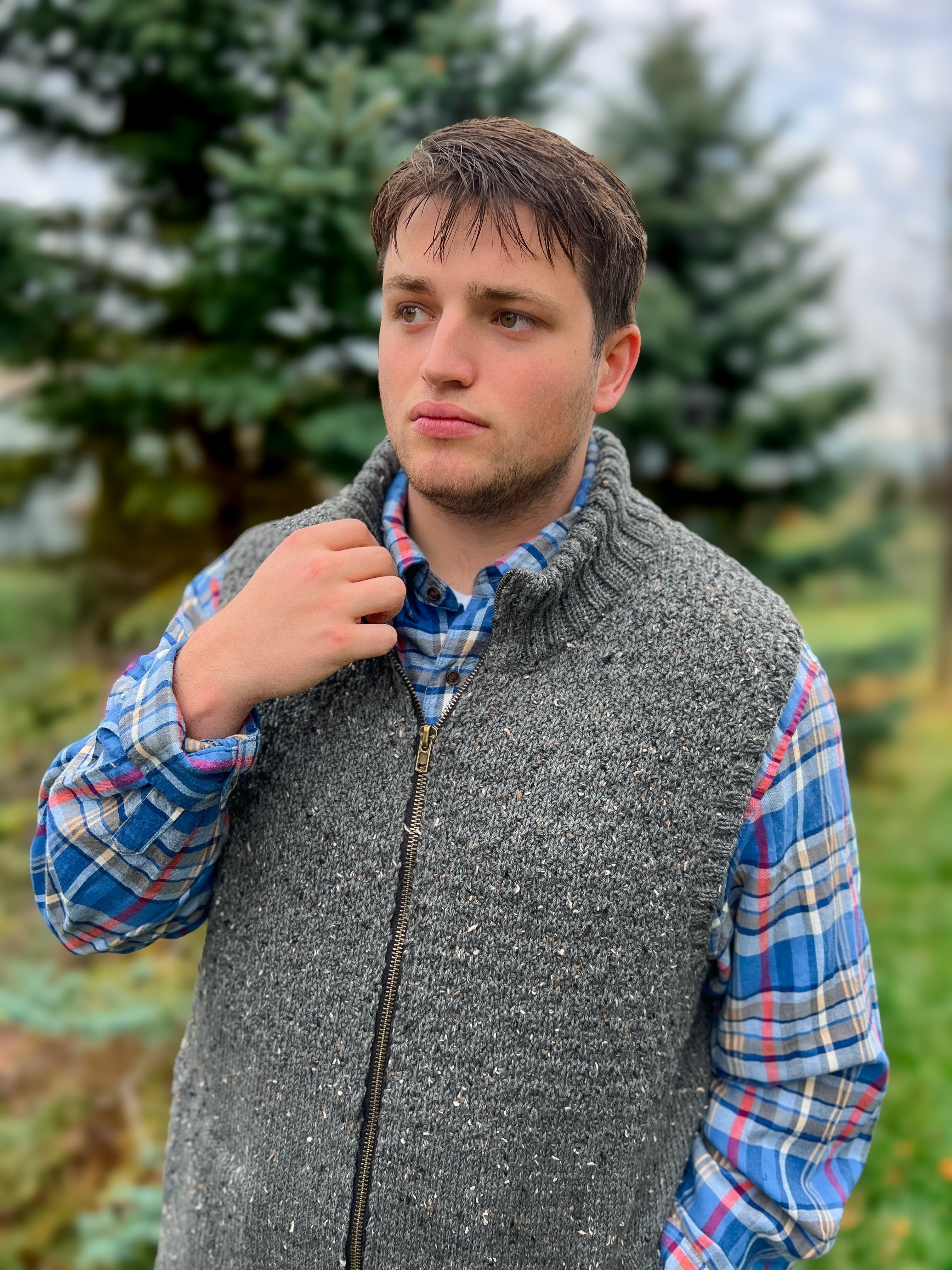 A young man stands outdoors, looking thoughtful, wearing the Harbor Ridge Knit Vest & Sweater Pattern in gray over a plaid shirt. Blurred pine trees and greenery form the background.