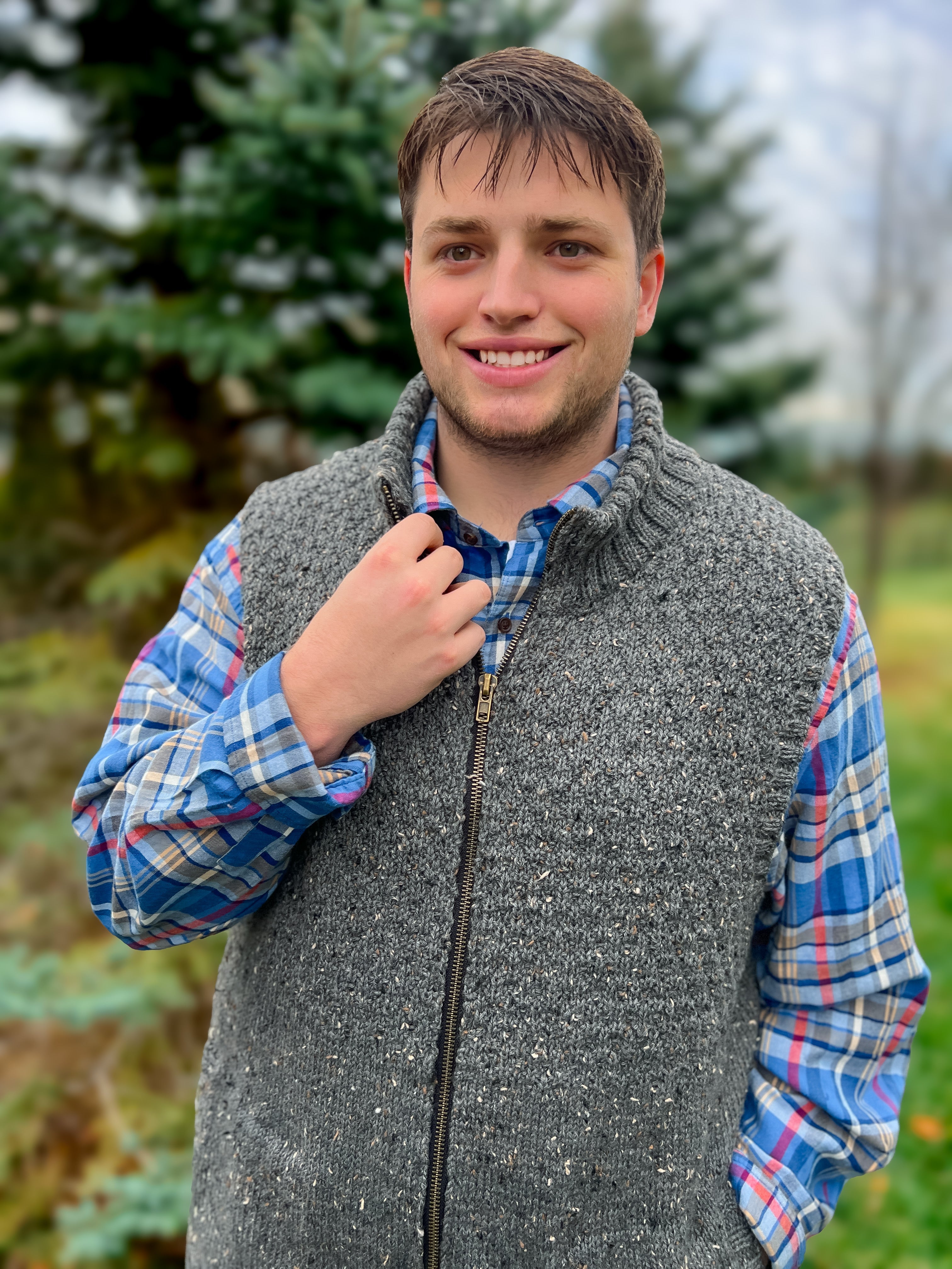 Wearing the Harbor Ridge Knit Vest in a classic seersucker stitch sweater pattern, a young man stands outside, smiling in front of evergreen trees and blurred greenery, his gray zip-front vest layered over a blue plaid shirt.