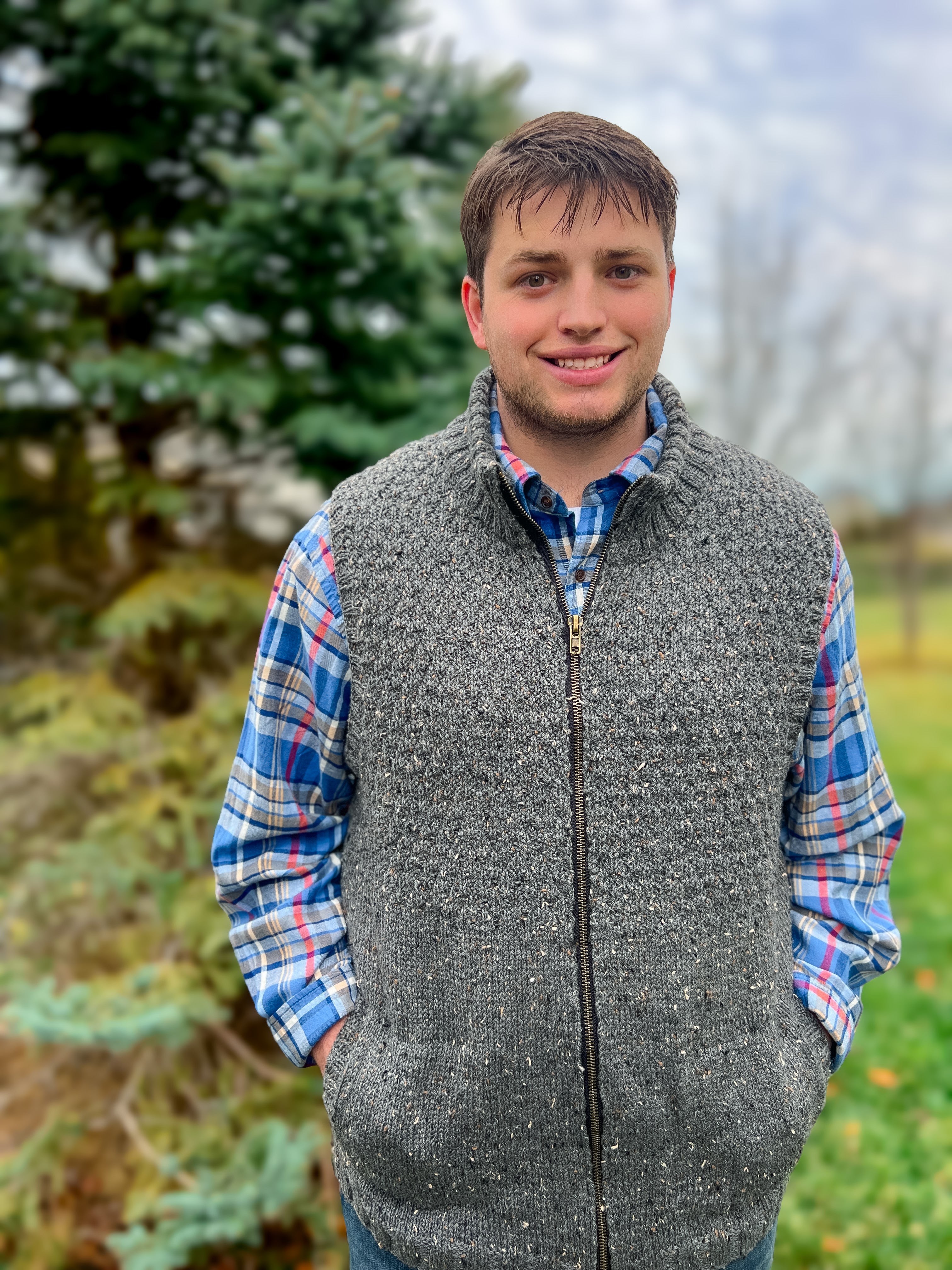 A young man stands outdoors, smiling, with his hands in his pockets, wearing the Harbor Ridge Knit Vest & Sweater Pattern in gray over a blue and red plaid shirt. A pine tree and grass can be seen in the background.