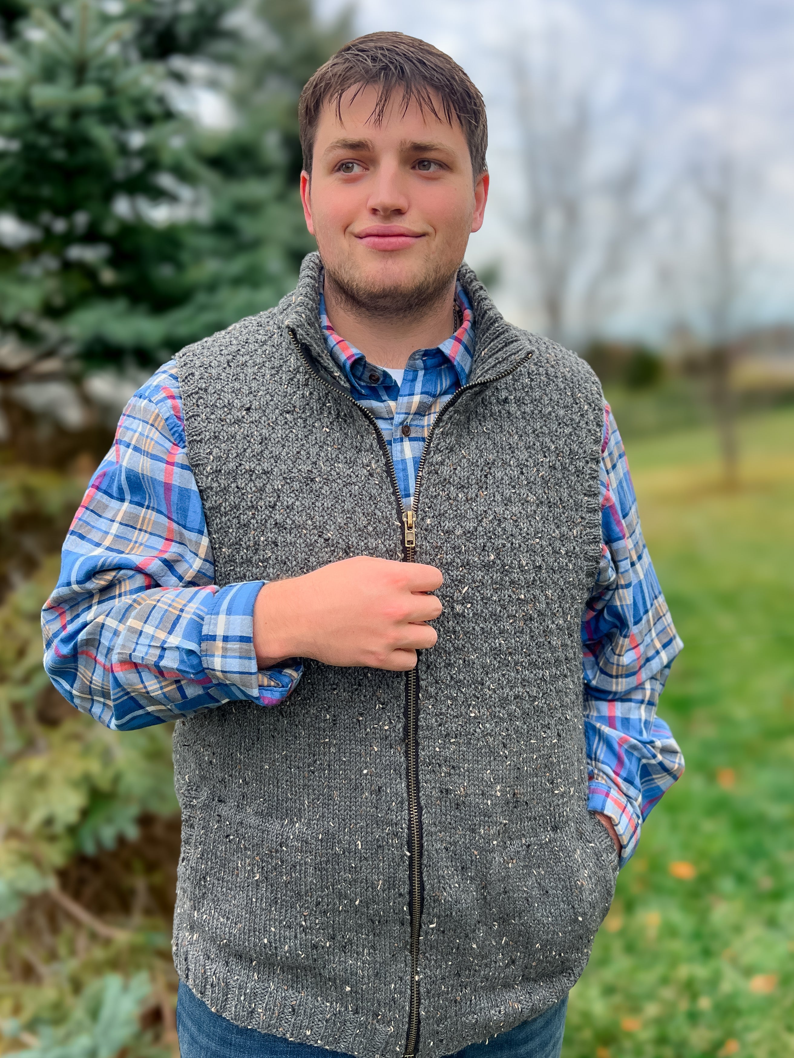 A man stands outdoors on grass, wearing the Harbor Ridge Knit Vest over a blue plaid shirt. He looks to the side with a slight smile, with evergreen trees and a cloudy sky softly blurred in the background.