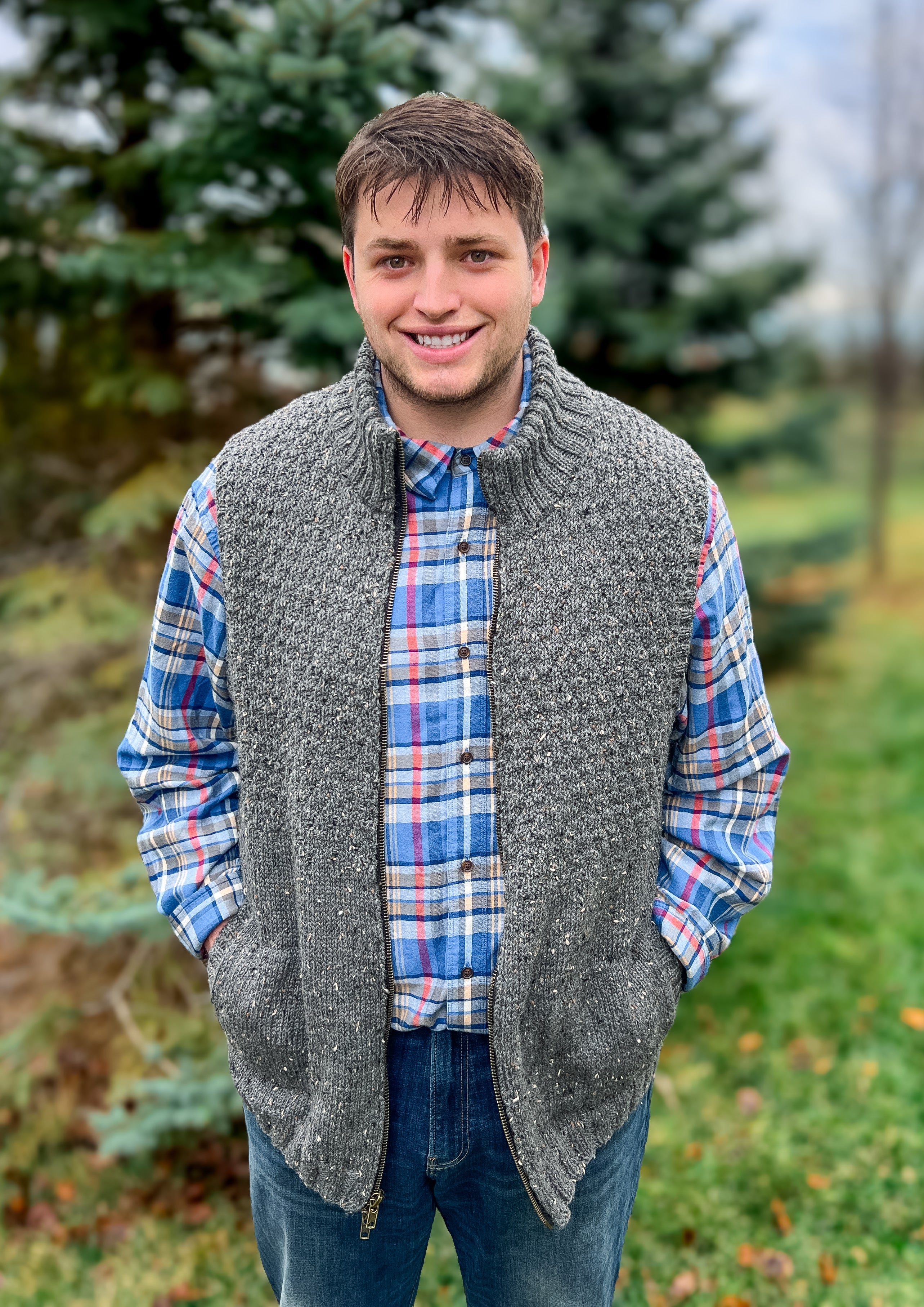 A young man stands outside, smiling with his hands in his pockets, wearing a gray Harbor Ridge Knit Vest & Sweater Pattern over a blue plaid shirt. Evergreen trees and green grass create a softly blurred background.