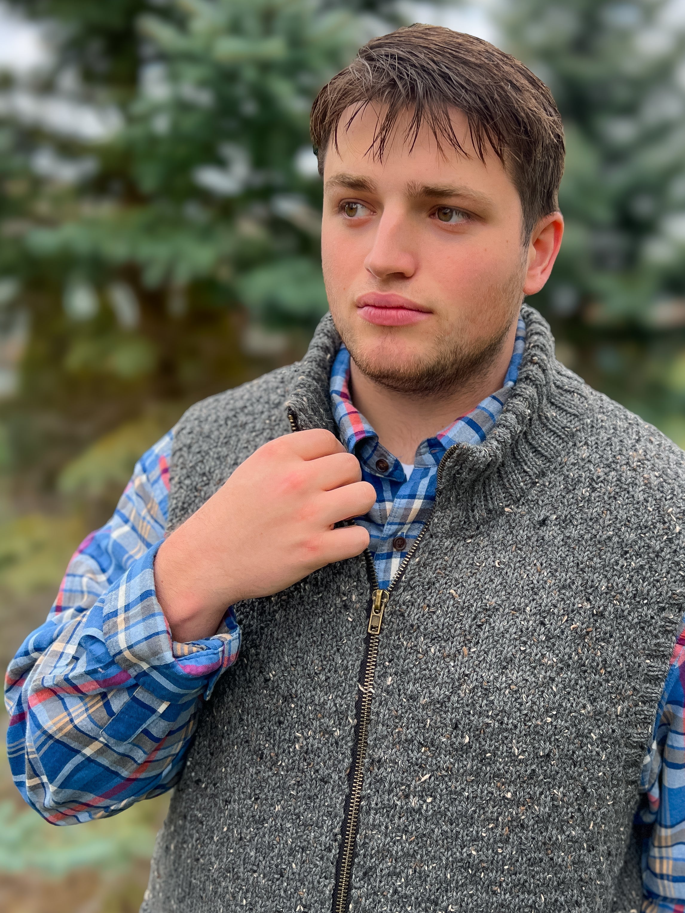 A young man stands outdoors amid blurred trees and greenery, holding his collar with one hand while wearing the Harbor Ridge Knit Vest & Sweater over a blue plaid shirt.