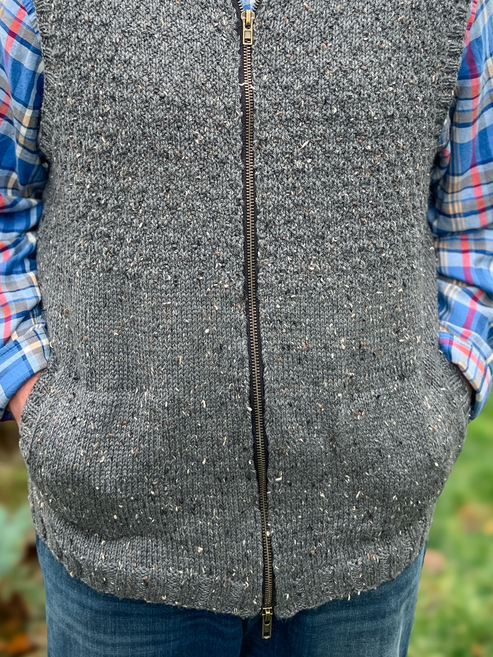 A person wearing the Harbor Ridge Knit Vest & Sweater—a chunky gray zip-front vest with pockets—over a blue and red plaid shirt, stands outdoors with hands tucked in the vest pockets. Green foliage blurs in the background, accentuating the textured knitting pattern of this cozy layer.