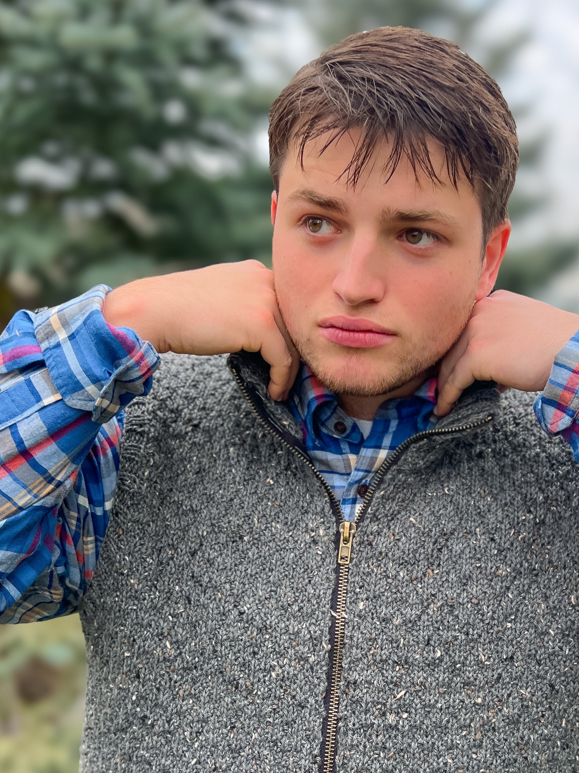 A young man stands outdoors among blurred evergreens, hands to his neck, wearing a blue plaid shirt topped with the Harbor Ridge Knit Vest, which features a subtle knitting pattern that brings texture and warmth to his look.