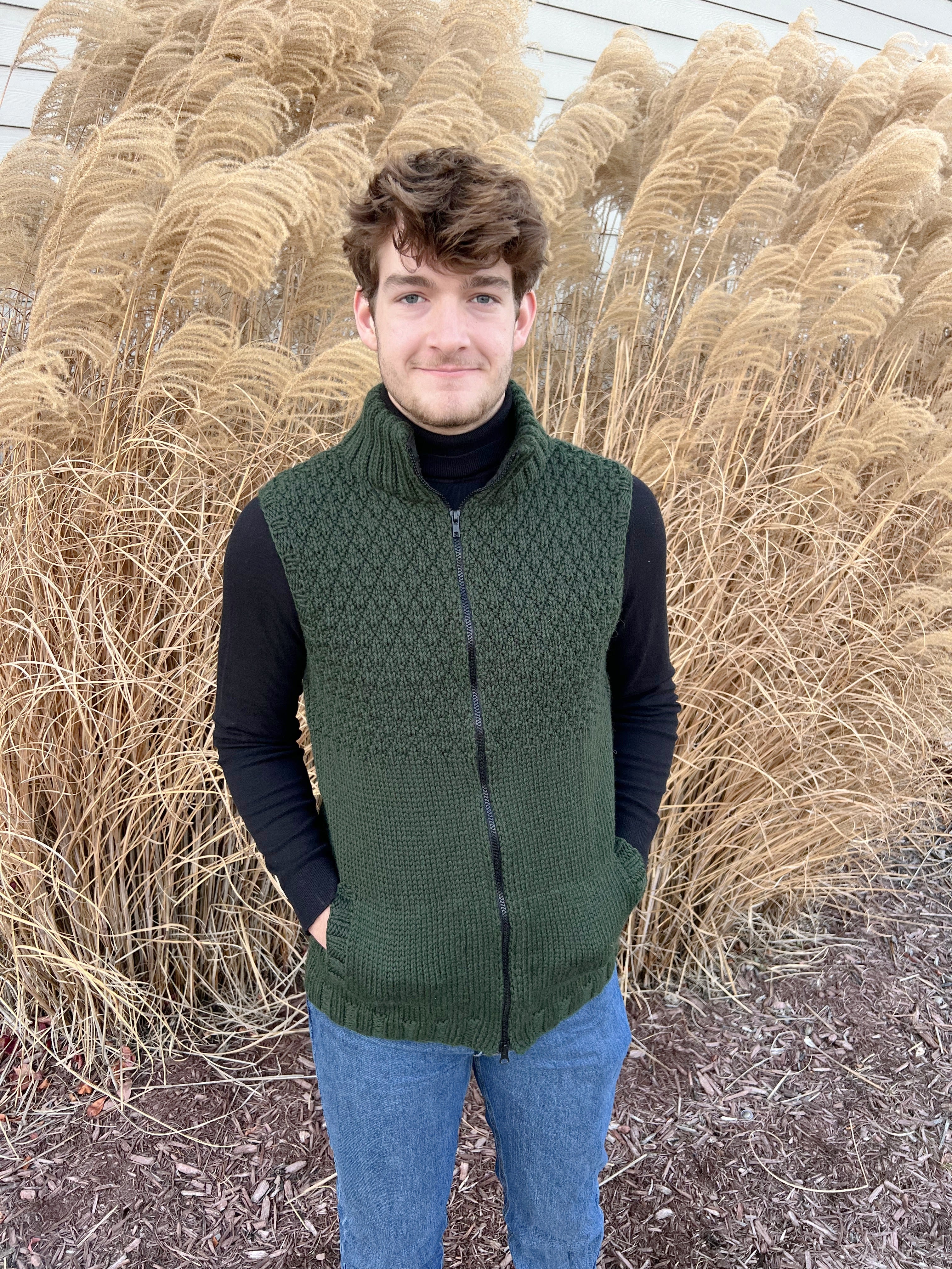 A young man stands in front of tall, golden ornamental grasses, smiling with his hands in his pockets while wearing the Harvey Knit Vest Pattern in green over a black shirt and blue jeans.