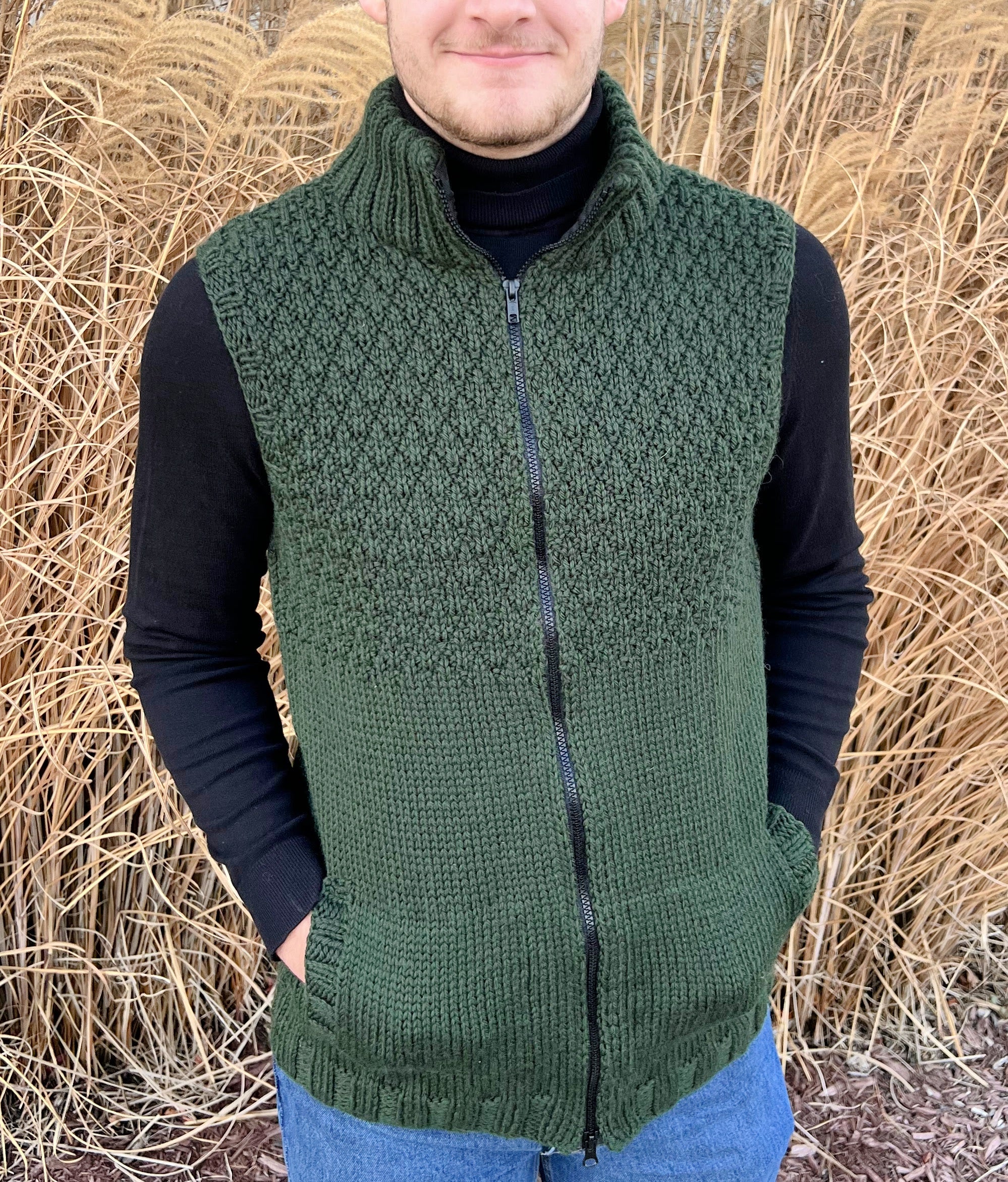 Wearing the Harvey Knit Vest Pattern in dark green, crafted with a seersucker stitch and zipped at the front over a black long-sleeve shirt, a person stands amid tall, dry grasses.