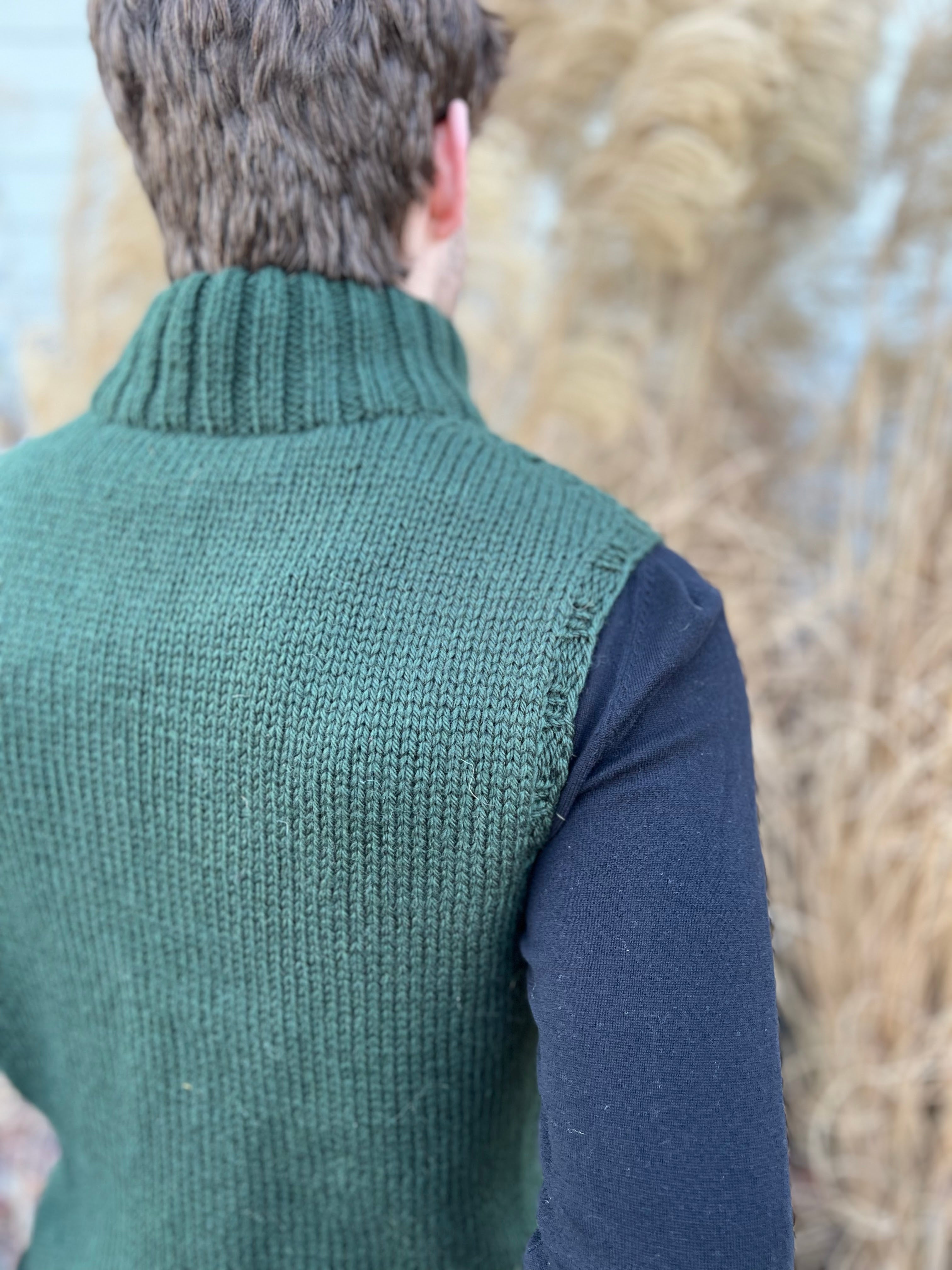 A person with short brown hair, seen from behind, wears a sleeveless dark green turtleneck vest knit in bulky yarn—the Harvey Knit Vest Pattern—over a black long-sleeve shirt. Tall dry grass is blurred in the background.