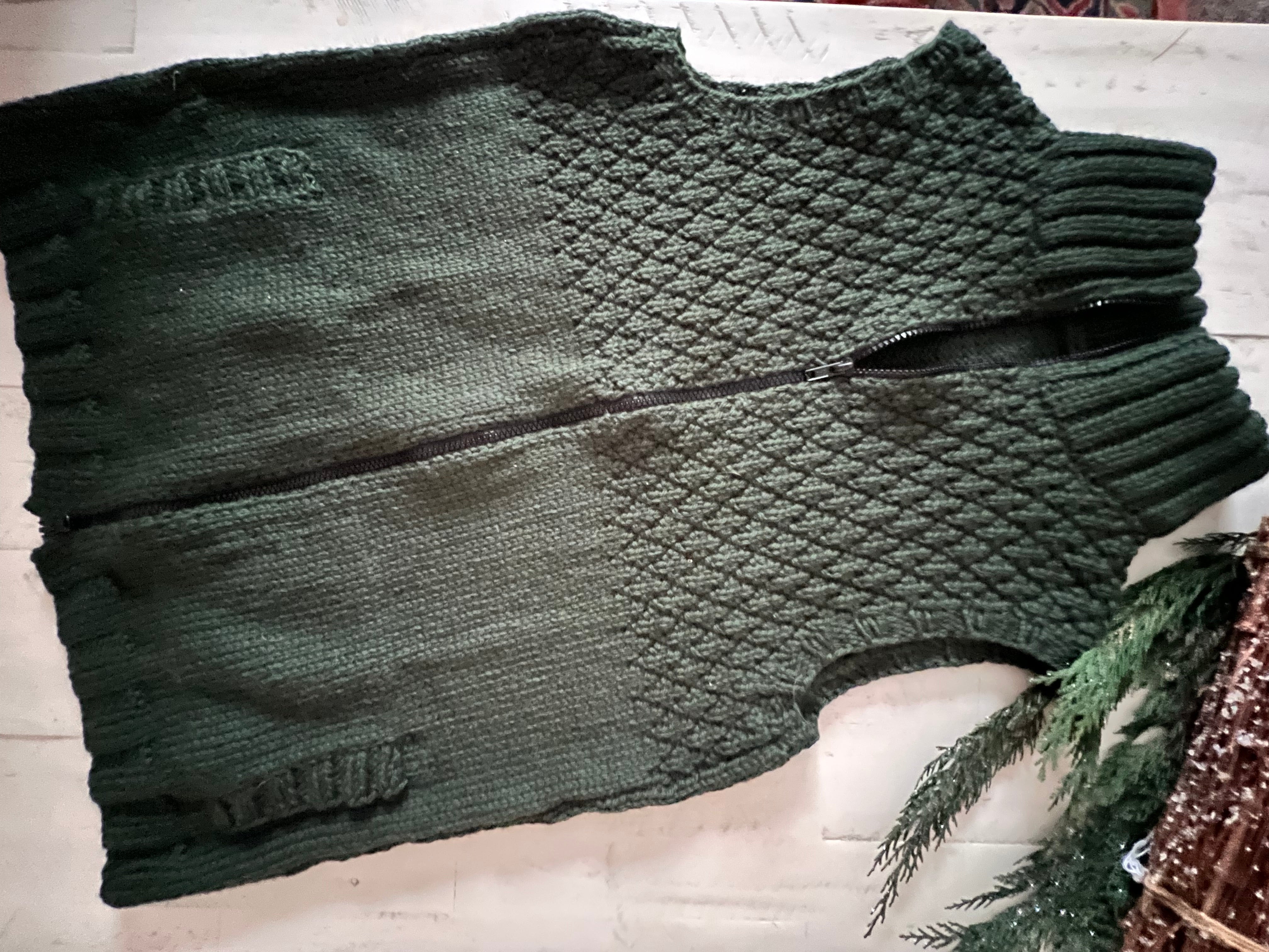 The Harvey Knit Vest Pattern, shown in a dark green zip-front design with a textured diamond pattern worked in seersucker stitch on the top half and smooth knitting on the bottom, is laid flat on a white surface. Pine branches appear in the bottom right corner.