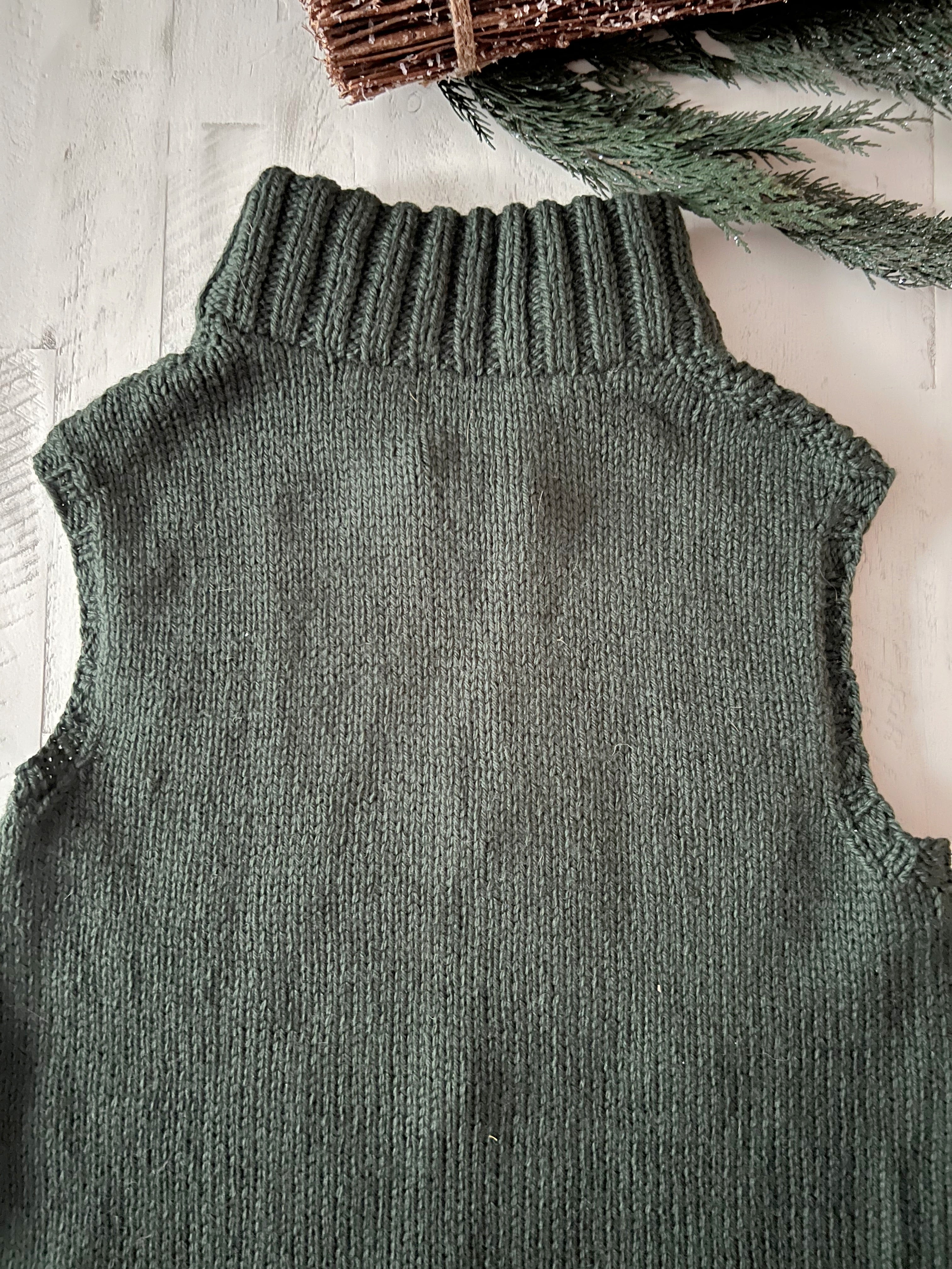 The Harvey Knit Vest Pattern is displayed laid flat on a white wooden surface, with green pine branches accenting the corner. This sleeveless, dark green sweater features a high, ribbed turtleneck and showcases textured elegance with its cozy bulky yarn and intricate seersucker stitch detailing—making it a stylish addition to any winter wardrobe.