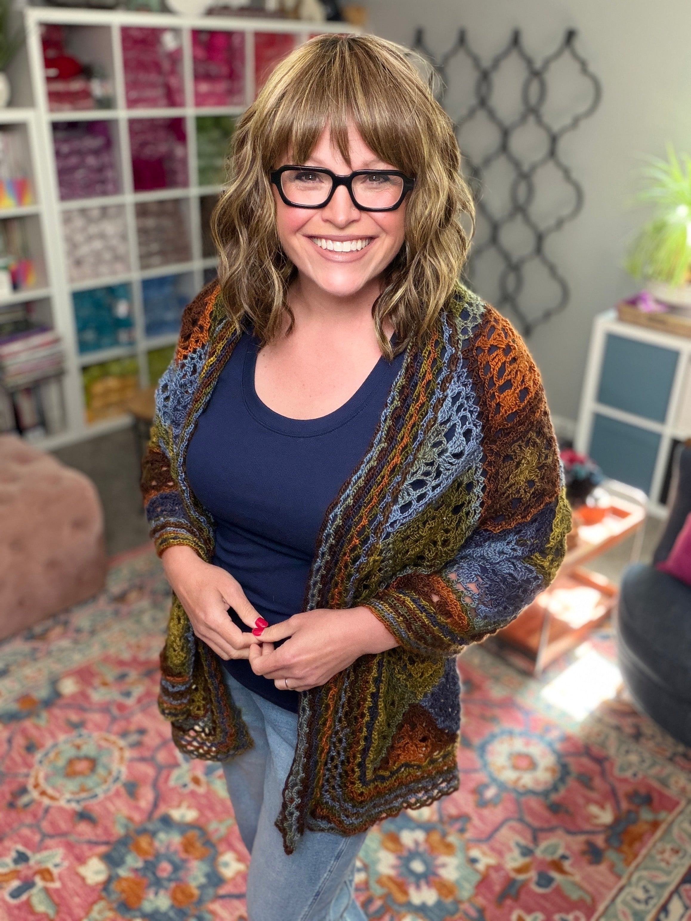 A smiling woman with wavy light brown hair, glasses, a navy shirt, and a colorful crocheted cardigan stands in a cozy room of yarn shelves—she's working on the Enchanted Crochet Motif Shawl Pattern, perfect for beginners.
