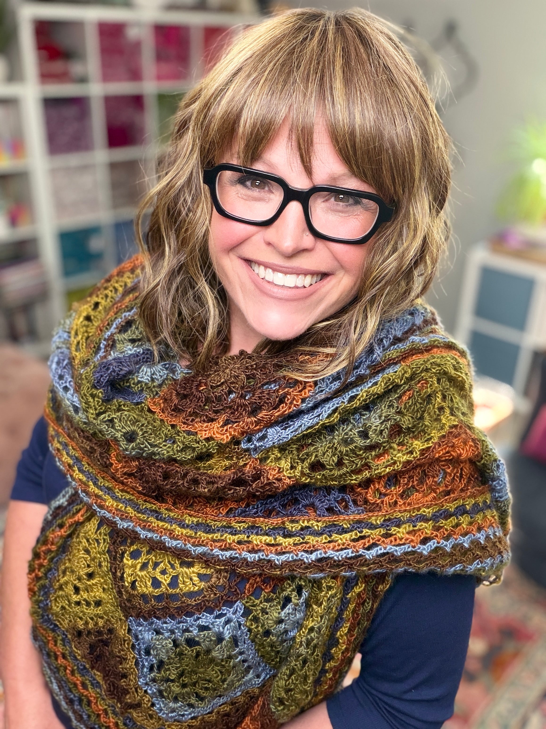 A smiling woman with wavy blonde hair and glasses wears a colorful shawl made from the Enchanted Crochet Motif Shawl Pattern—featuring blue, green, yellow, and brown squares—standing indoors in a cozy, well-lit room.