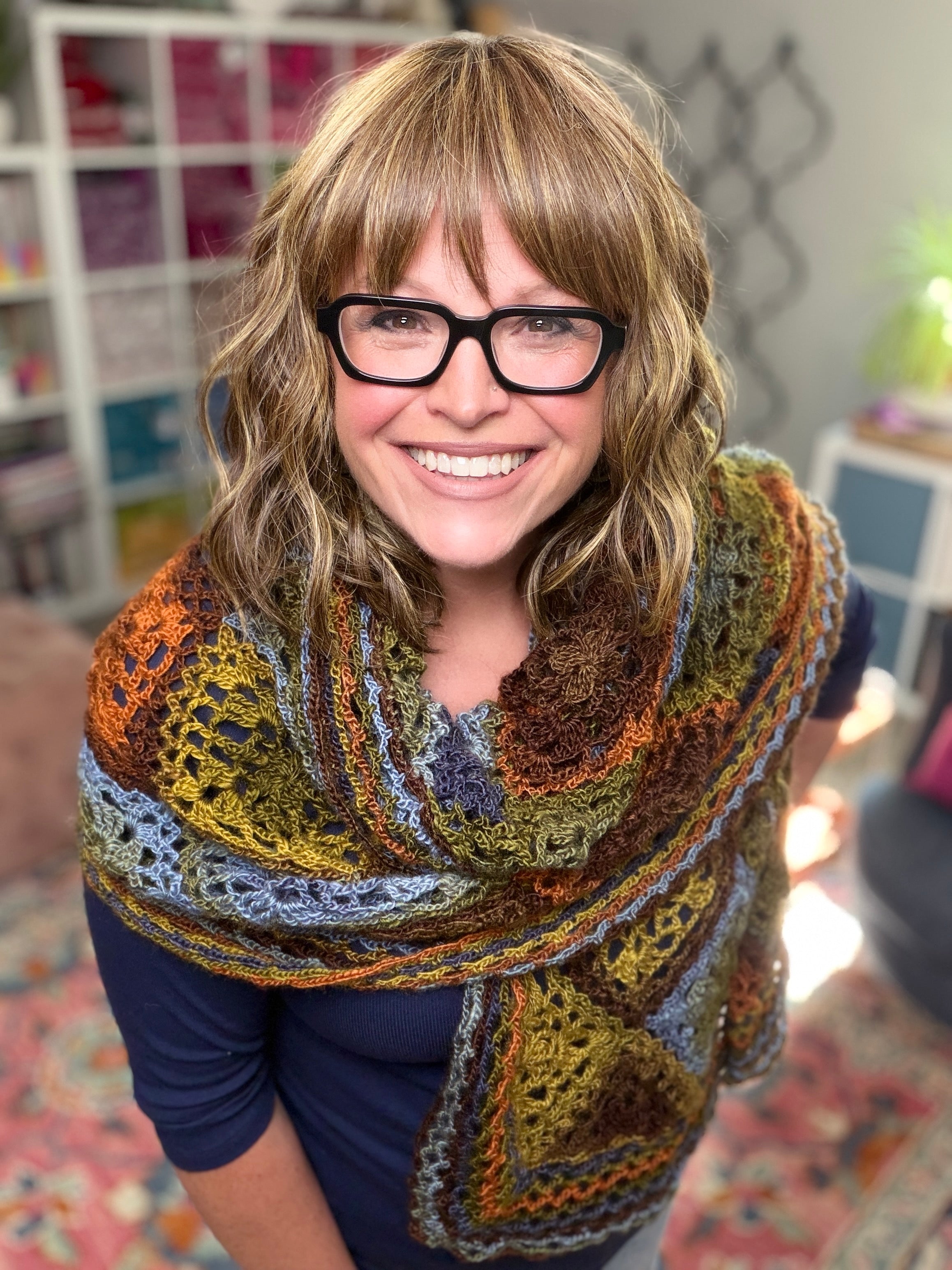 A smiling woman with wavy, light brown hair and black glasses wears a colorful shawl made from the Enchanted Crochet Motif Shawl Pattern over a navy top, standing indoors with shelves and a patterned rug in the background.