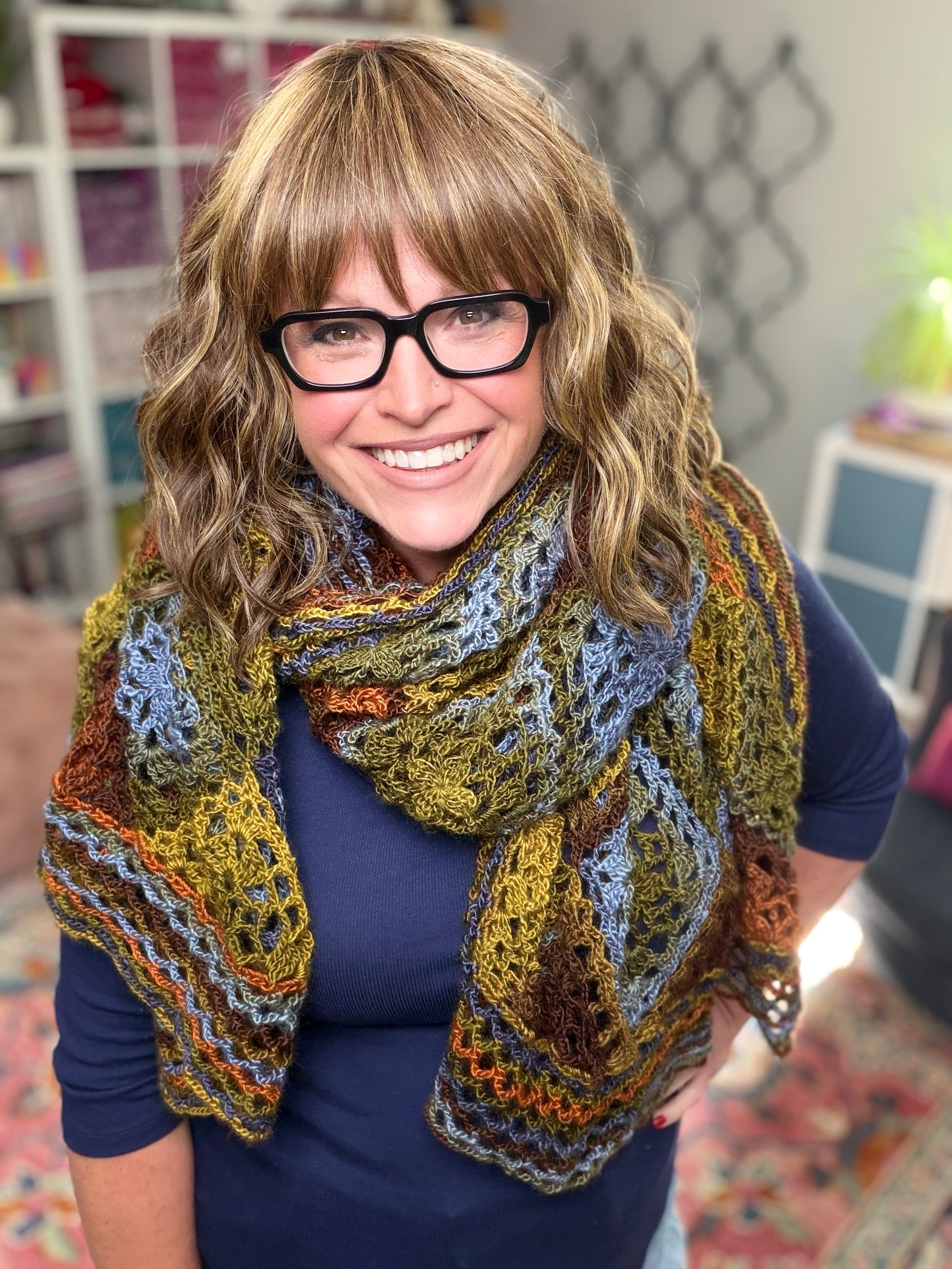 A smiling woman with glasses and wavy, shoulder-length hair stands indoors, wrapped in a blue, green, and brown shawl made from the Enchanted Crochet Motif Shawl Pattern. Shelves and a patterned carpet can be seen in the background.