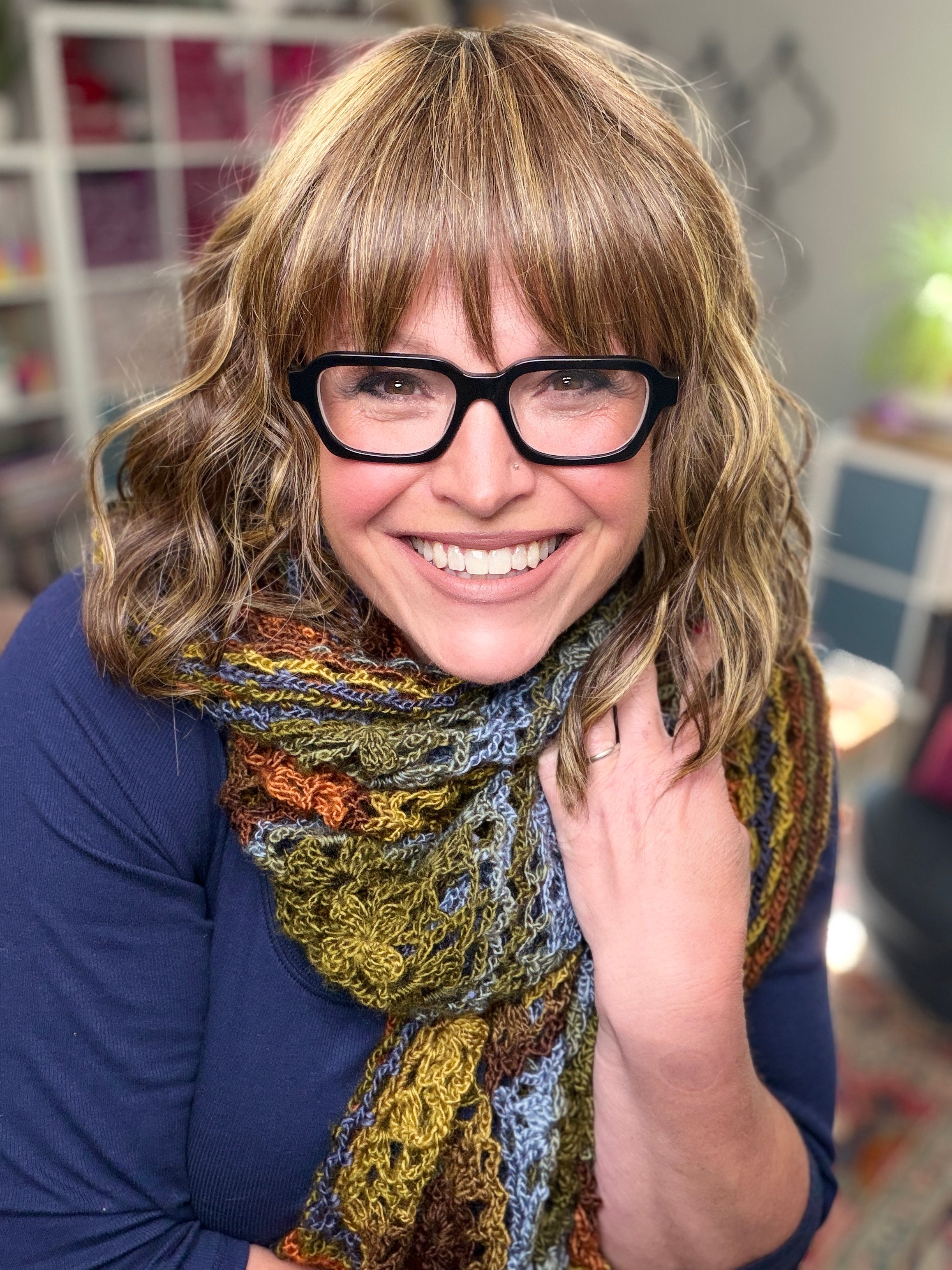 A smiling woman with wavy light brown hair and black glasses wears a multicolored scarf made from the Enchanted Crochet Motif Shawl Pattern, paired with a navy blue top. She poses indoors, hand on scarf; blurred shelves and plants in the background.
