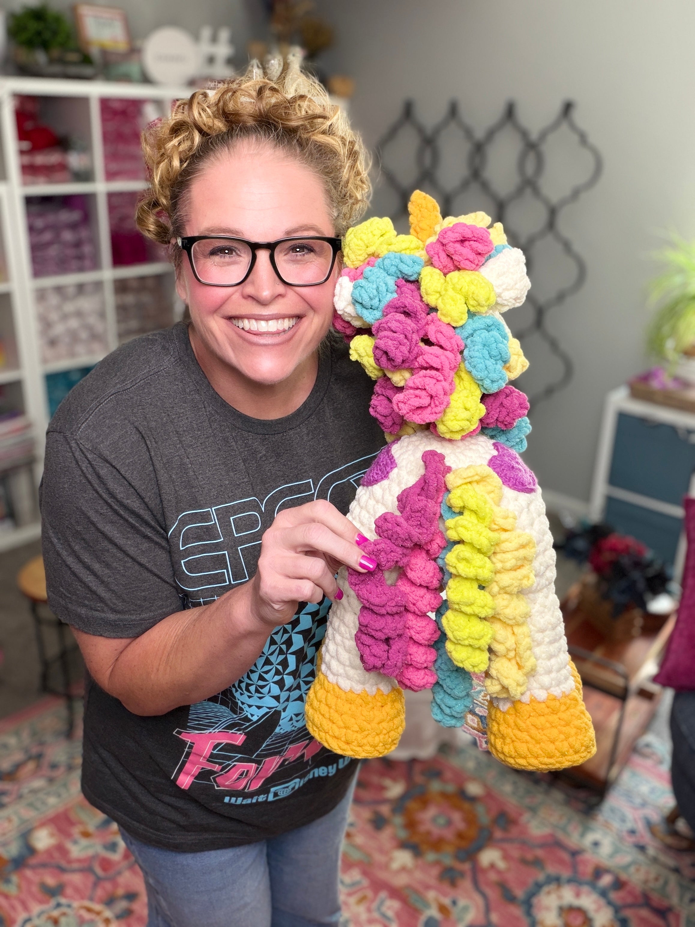 A smiling woman with curly blonde hair and glasses holds a large unicorn made from the Sparkle Crochet Unicorn Amigurumi Pattern, standing in a cozy, yarn-filled room that highlights her latest crocheted creation.