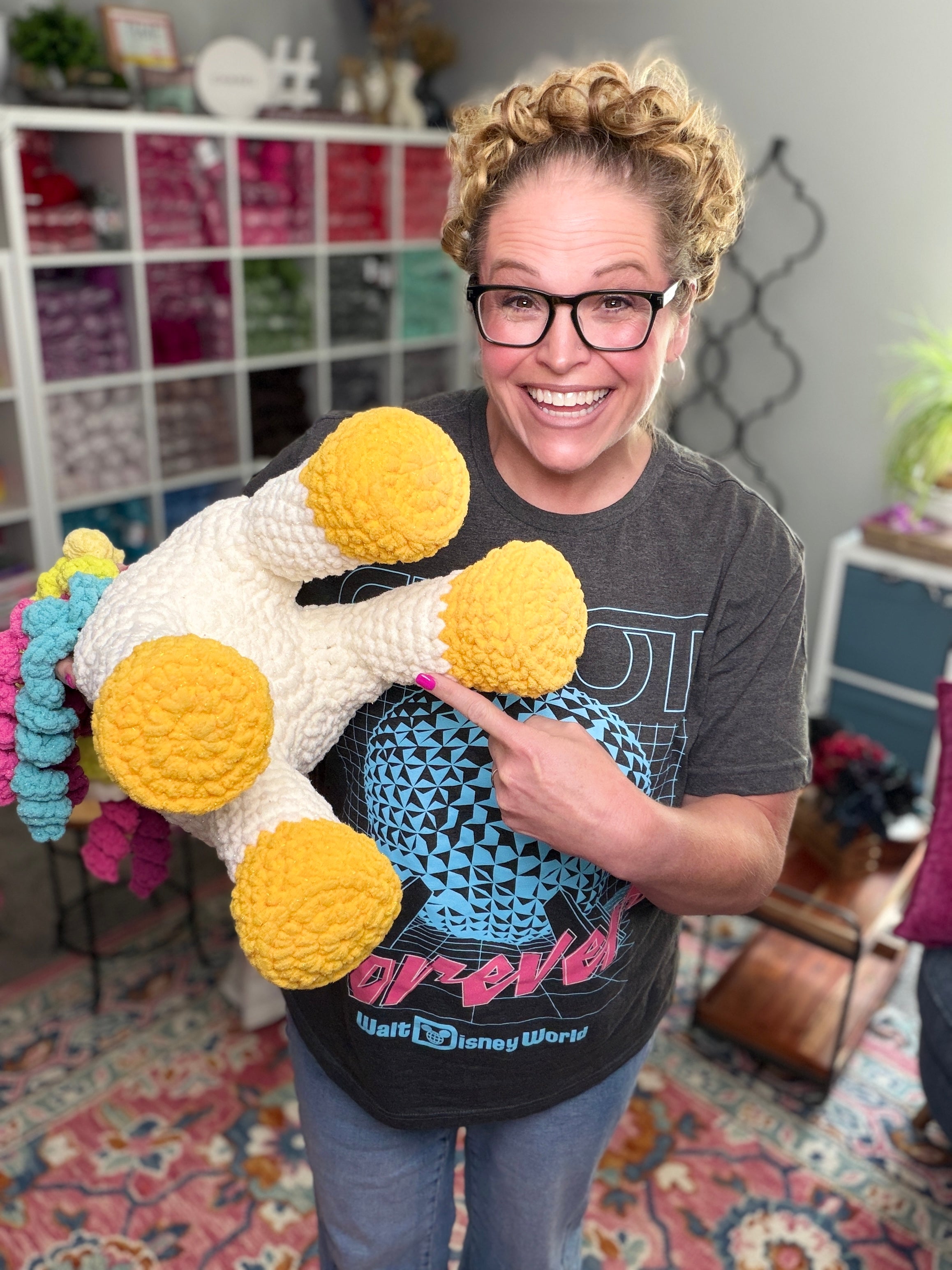 A smiling woman with glasses and curly hair holds a large Sparkle Crochet Unicorn Amigurumi Pattern sample, standing in a colorful craft room with yarn shelves in the background.