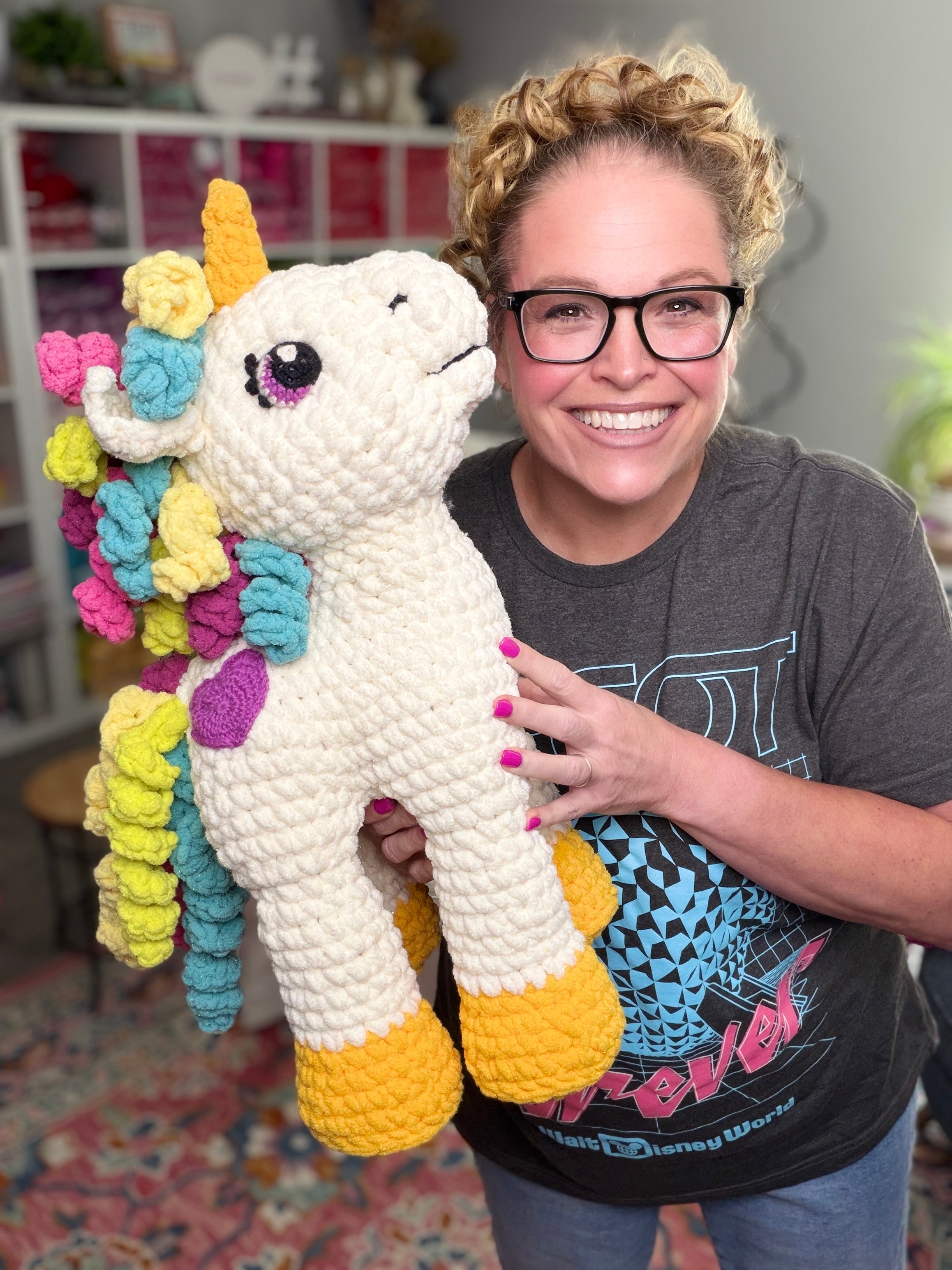 A smiling woman with glasses and curly hair holds a large unicorn plushie made from the Sparkle Crochet Unicorn Amigurumi Pattern, in a cozy room with shelves and a patterned rug in the background.