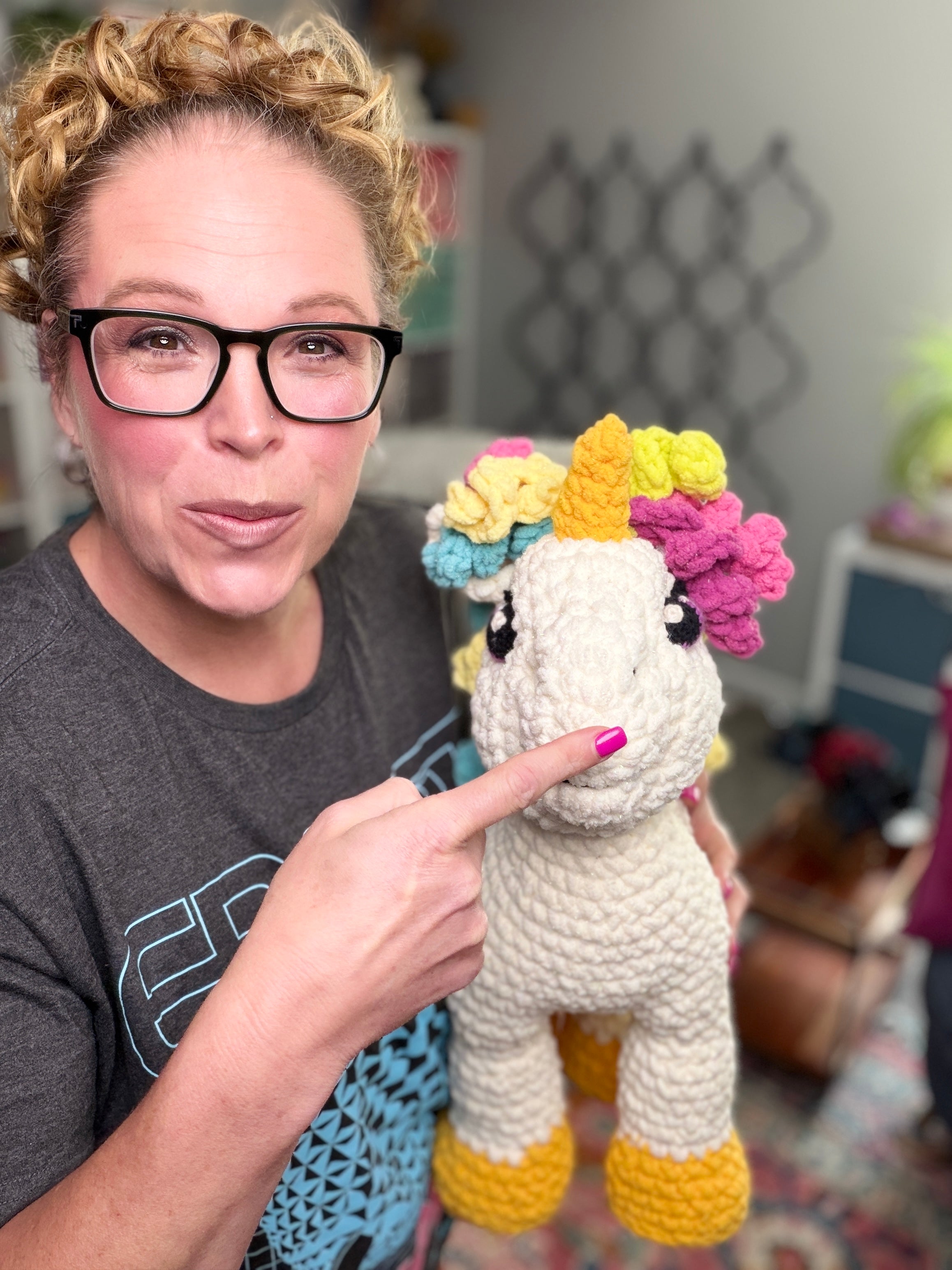 A woman with glasses and curly hair smiles as she proudly displays her latest creation—the Sparkle Crochet Unicorn Amigurumi Pattern, featuring a colorful mane and yellow horn, in a cozy, well-lit room.