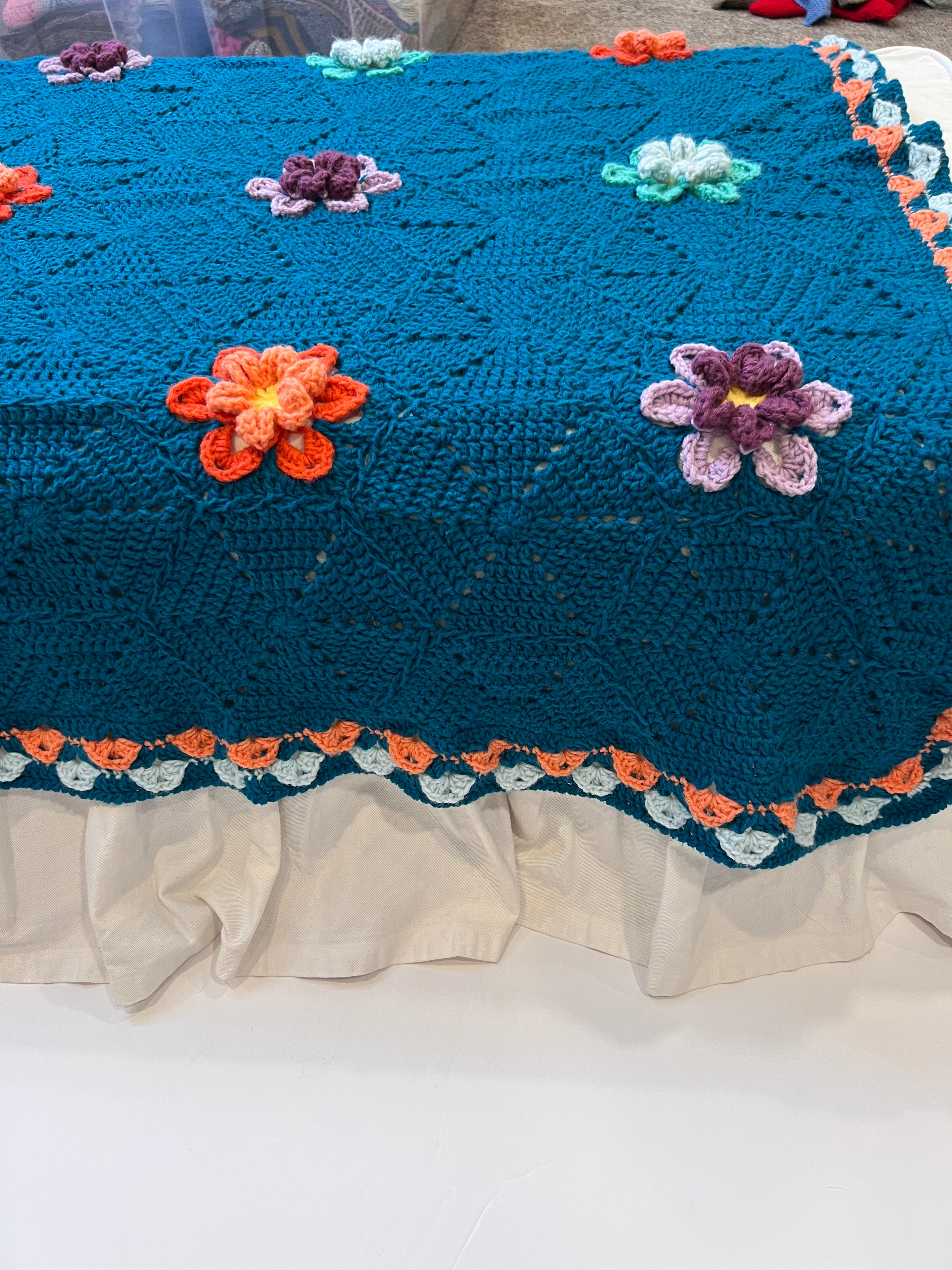 A blue Wildflower Hexagon Motif Crochet Blanket, featuring vibrant colorful 3D flower decorations and an orange and white border, is draped over a white fabric-covered surface.