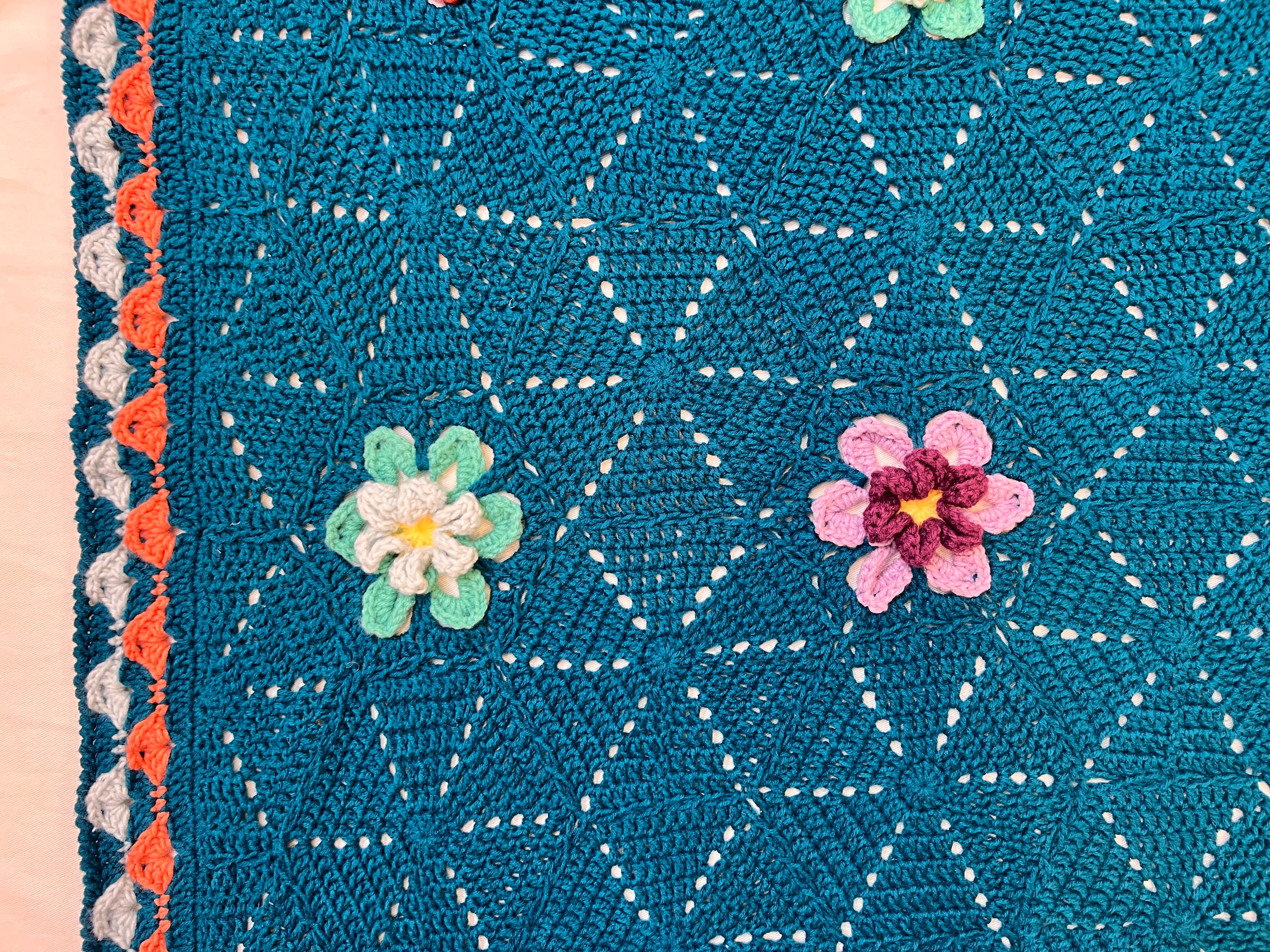 The Wildflower Hexagon Motif Crochet Blanket Pattern features a teal blanket adorned with geometric hexagon motifs and three-dimensional crocheted flowers in green, white, pink, and yellow. This design showcases a beautiful crochet motif pattern along with scalloped edging in coral, white, and gray.
